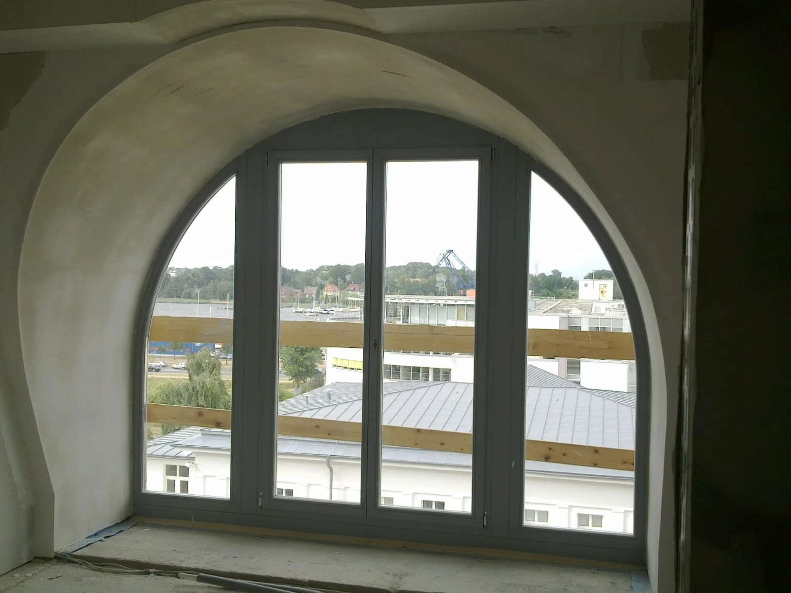 Ein Fenster in einem Raum unter einem Bogen, mit Holzstützen an den Seiten, Blick nach draußen auf Gebäude, Bäume, Wasser und eine Brücke in der Ferne.
