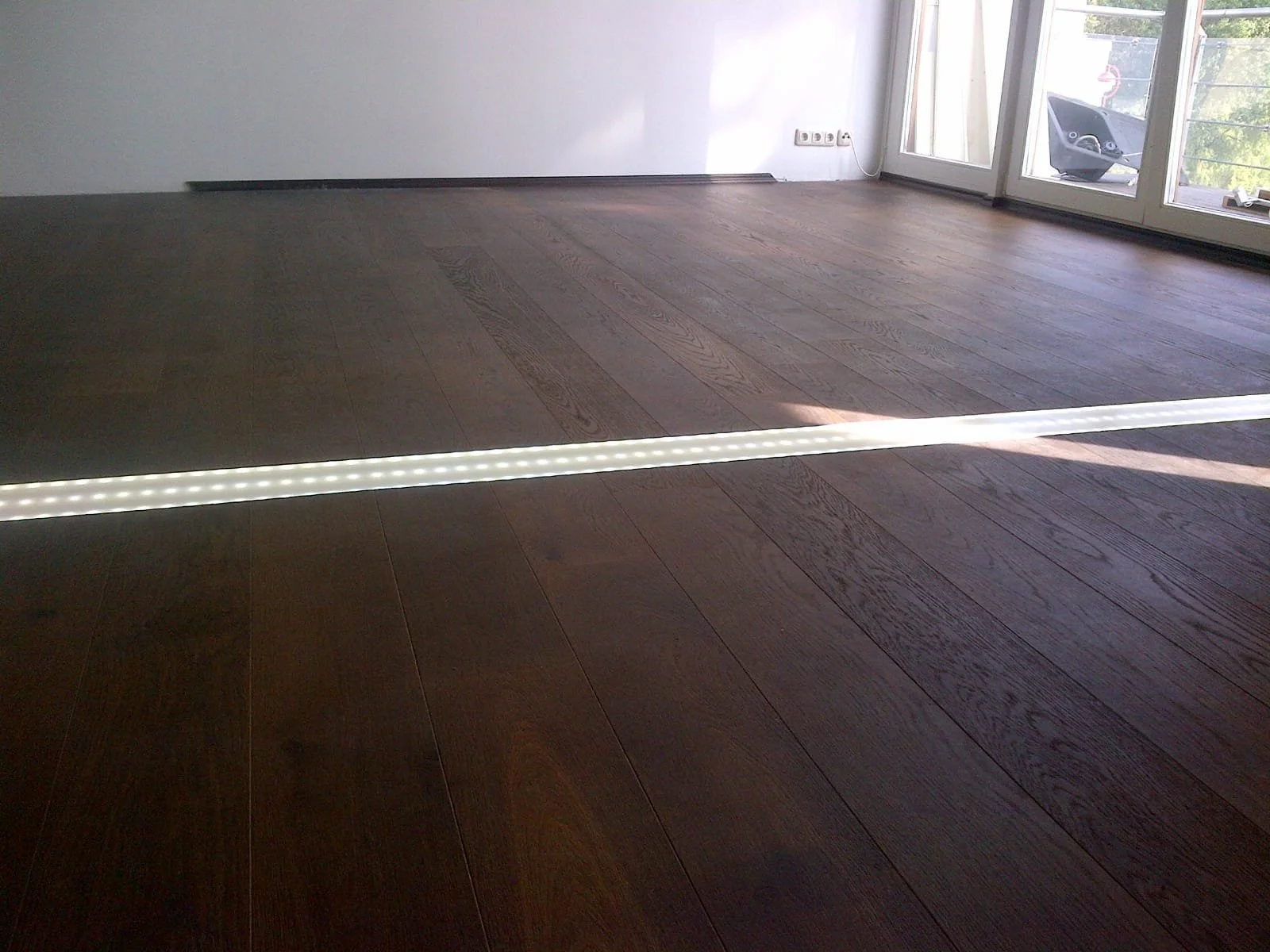 Ein leeres Zimmer mit dunklem Holzboden, einer weißen Wand, und großen Fenstern, durch die Sonnenlicht einfällt.