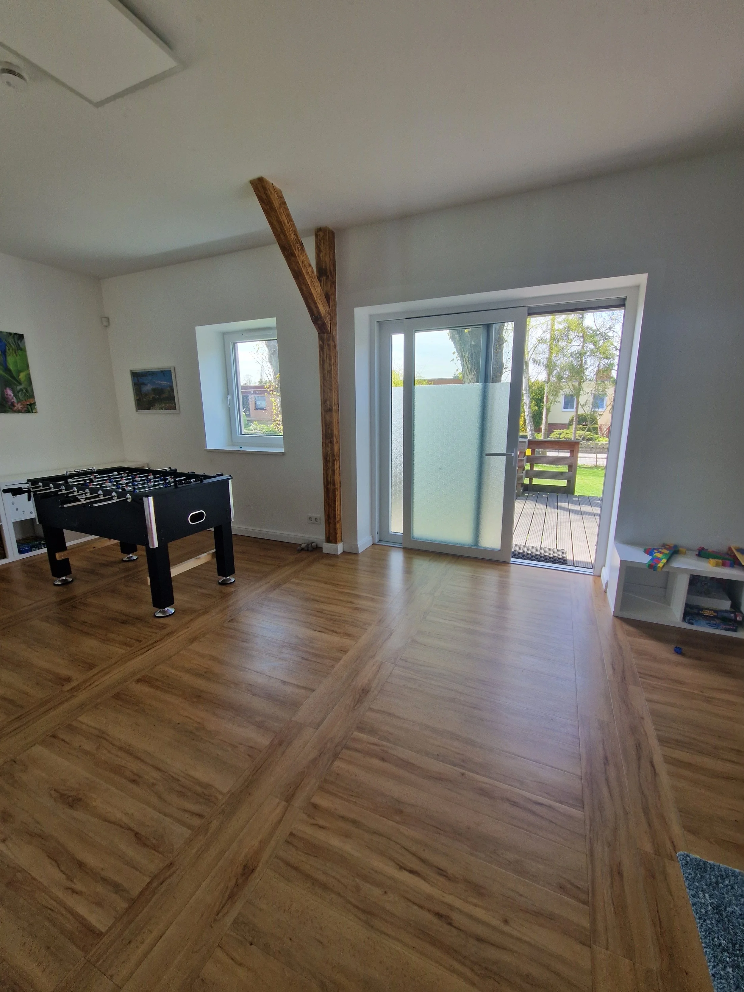 Innenraum eines Wohnzimmers mit Holzboden, einem Tür- und Fensterausblick nach draußen, einem Tischfussballspiel und einem kleinen Regal mit Spielzeugen.