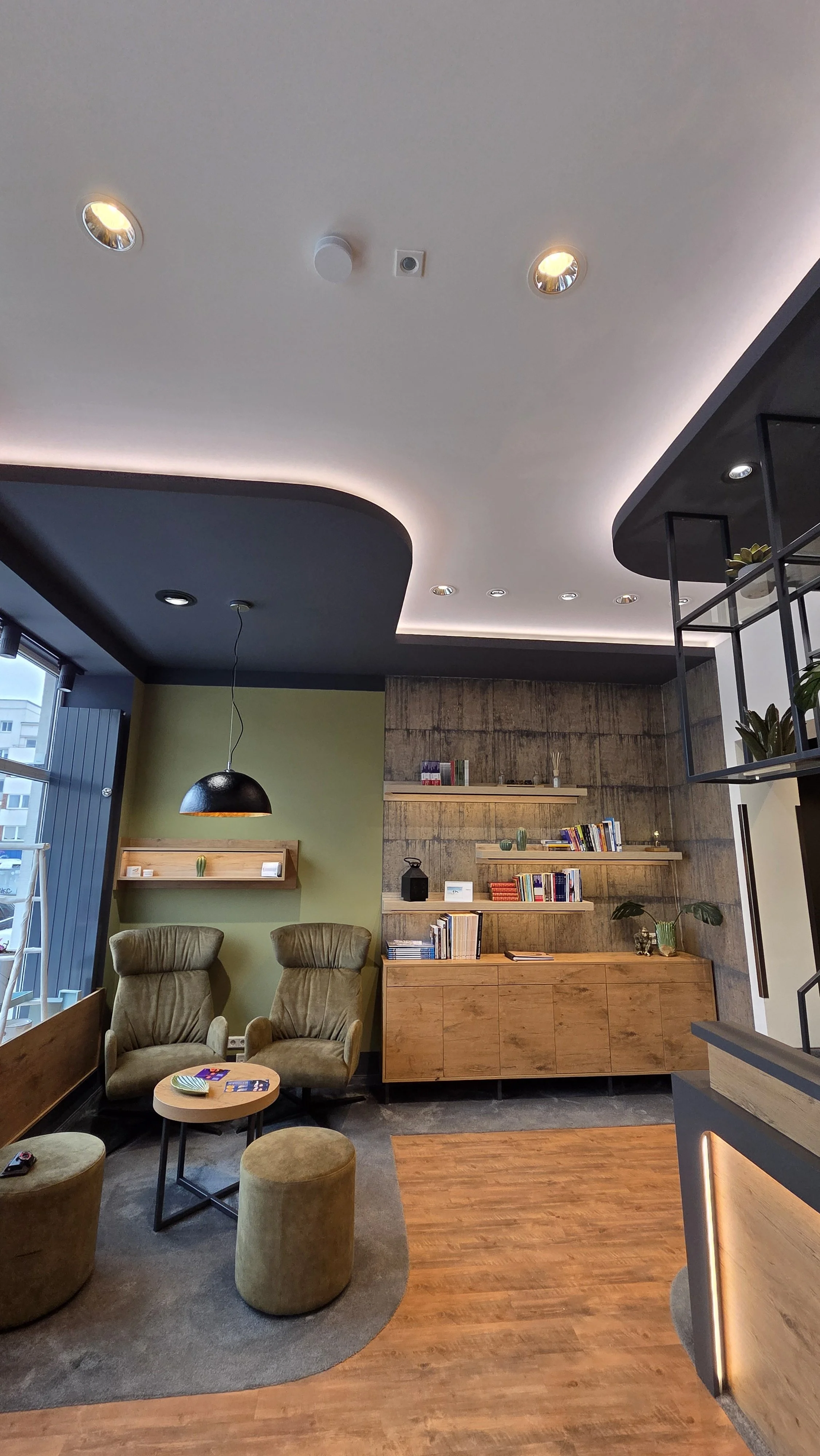 Modernes Wohnzimmer mit zwei bequemen Sessel, einem kleinen runden Tisch, einem Regal mit Büchern und Dekorationsgegenständen, grünen Pflanzen, einer dunklen Decke mit eingelassenen Lampen, und einer Wand mit Holzverkleidung.