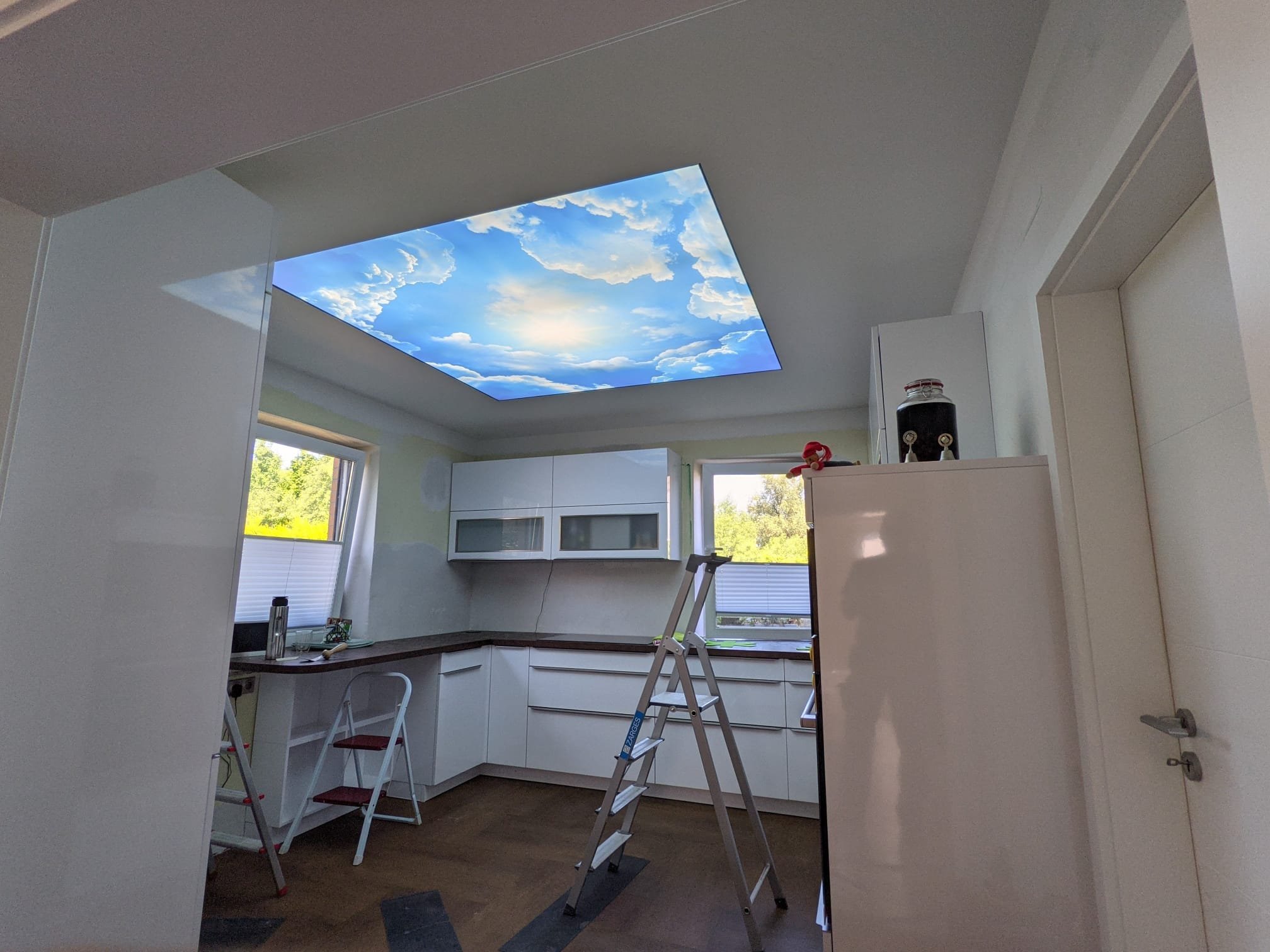 Innenaufnahme einer Küche mit weißem Schrank, einem Fenster, Arbeitsplatte und einem imaginären Himmel an der Decke.