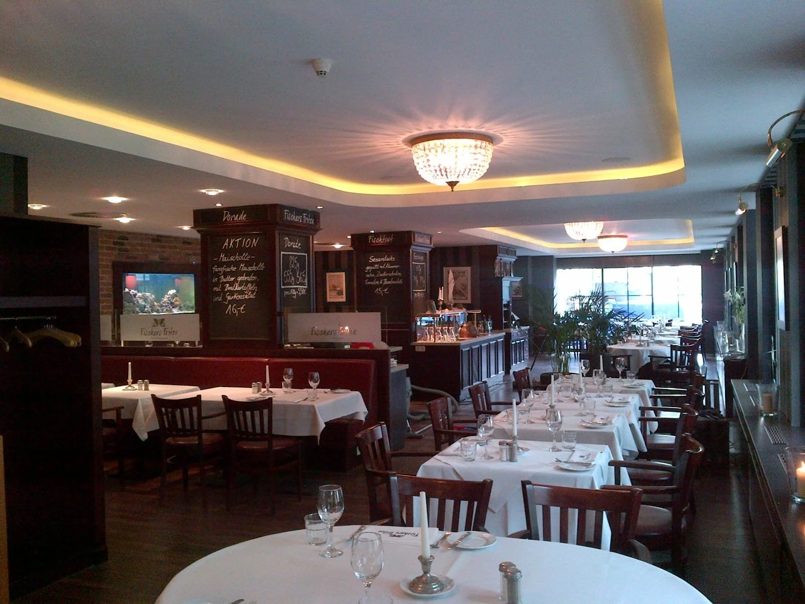 Innenansicht eines eleganten Restaurants mit weissen Tischen, Stühlen und Tischen mit Besteck, Gläsern und Kerzen, große Fenster im Hintergrund mit Sitzgelegenheiten und Pflanzen, an der Decke eine moderne Kronleuchter und indirekte Beleuchtung.
