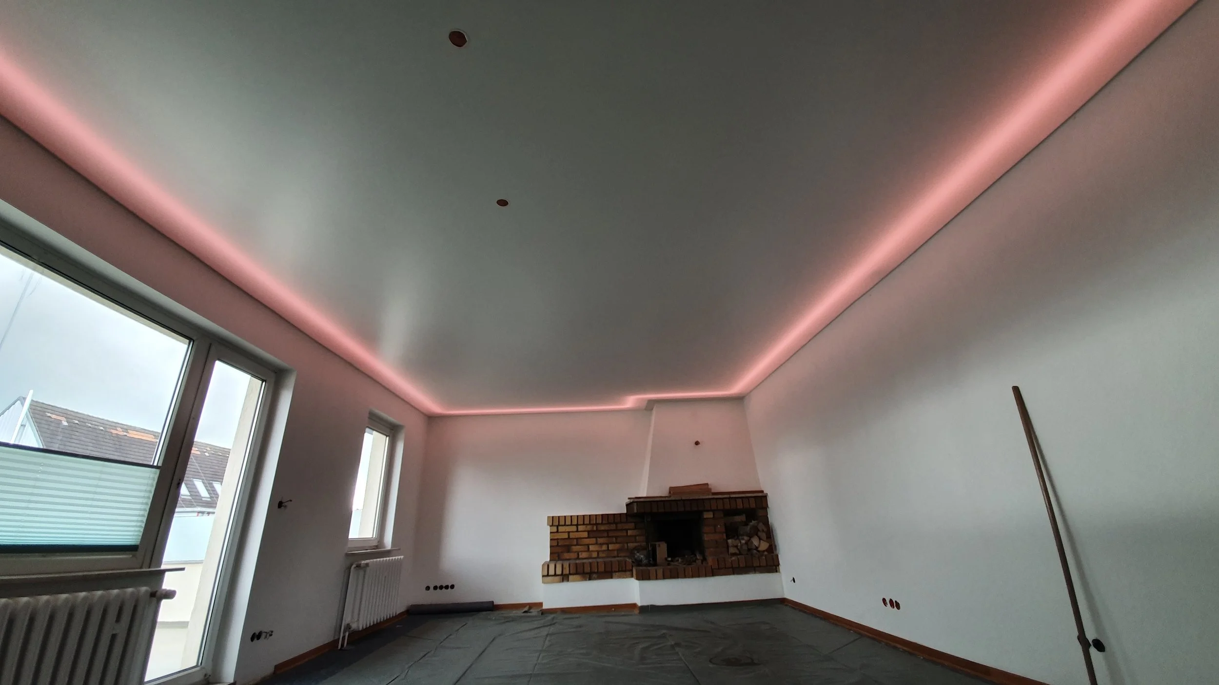 Innenraum eines Zimmers mit einer hohen Decke, LED-Beleuchtung entlang der Dachkante, große Fenster, einen Kamin, und eine unvollständige Einrichtung.