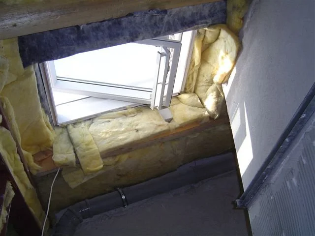 Ein offenes Dachfenster mit gelber Dämmwolle im Dachgeschoss, sichtbare Isolierung und Dachträger.