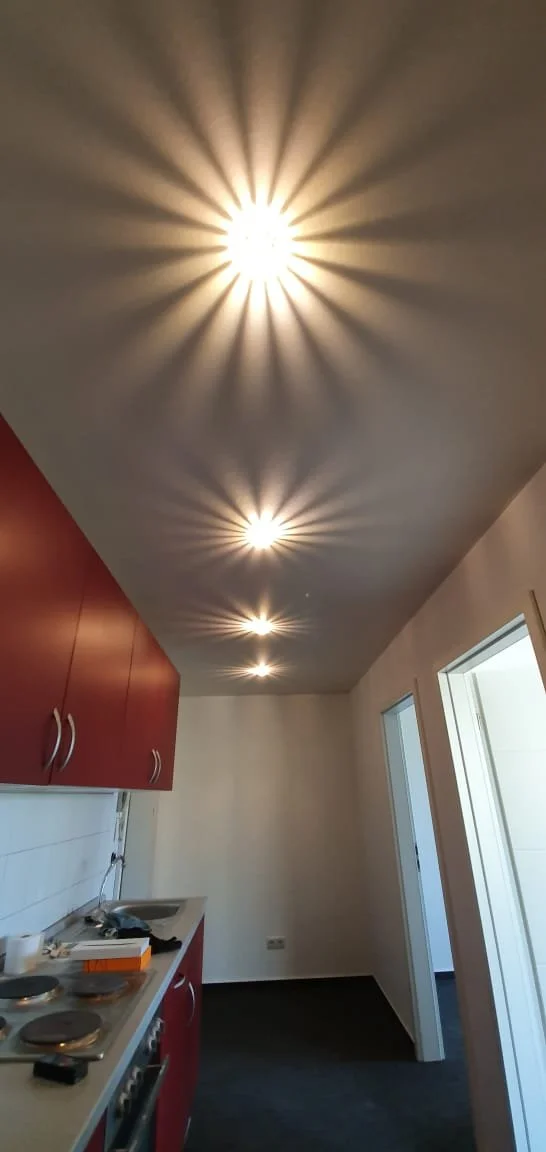 Innenraum einer modernen Küche mit roten Schränken und weißen Wänden, Deckenbeleuchtung mit Lichtmustern, die an Sonnenstrahlen erinnern, und einer Tür, die auf einen Balkon oder eine Terrasse führt.