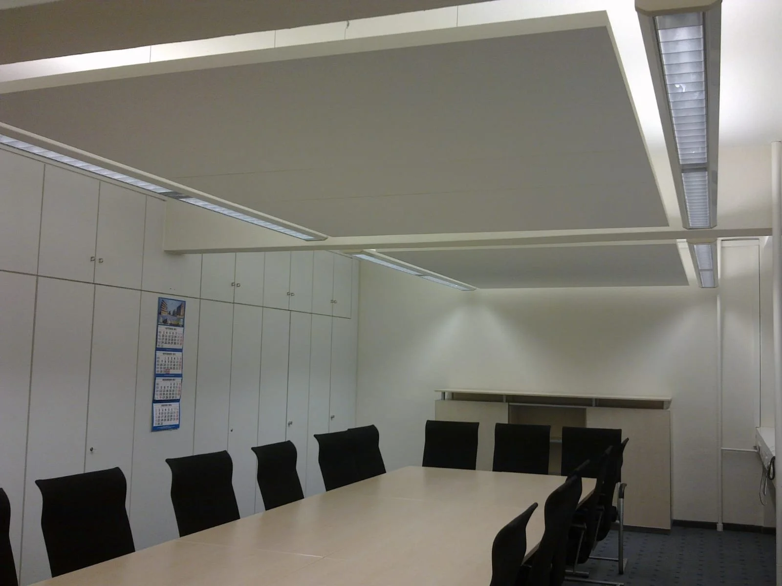 Ein Konferenzraum mit schwarzen Stühlen um einen rechteckigen Tisch, weiße Schränke an der Wand, eine Wandkalender und Deckenleuchten.