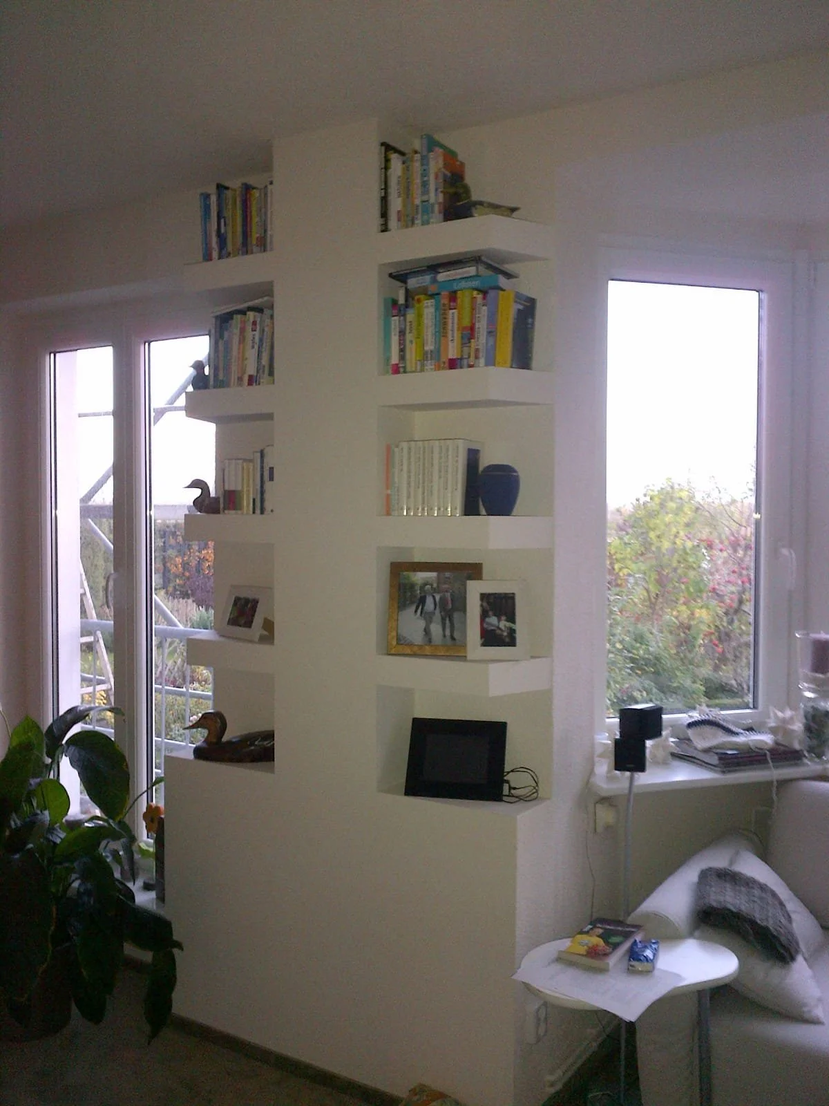 Innenaufnahme eines Wohnzimmers mit Bücherregal, zwei Fenster, einem Sofa und gefülltem Nachttisch.