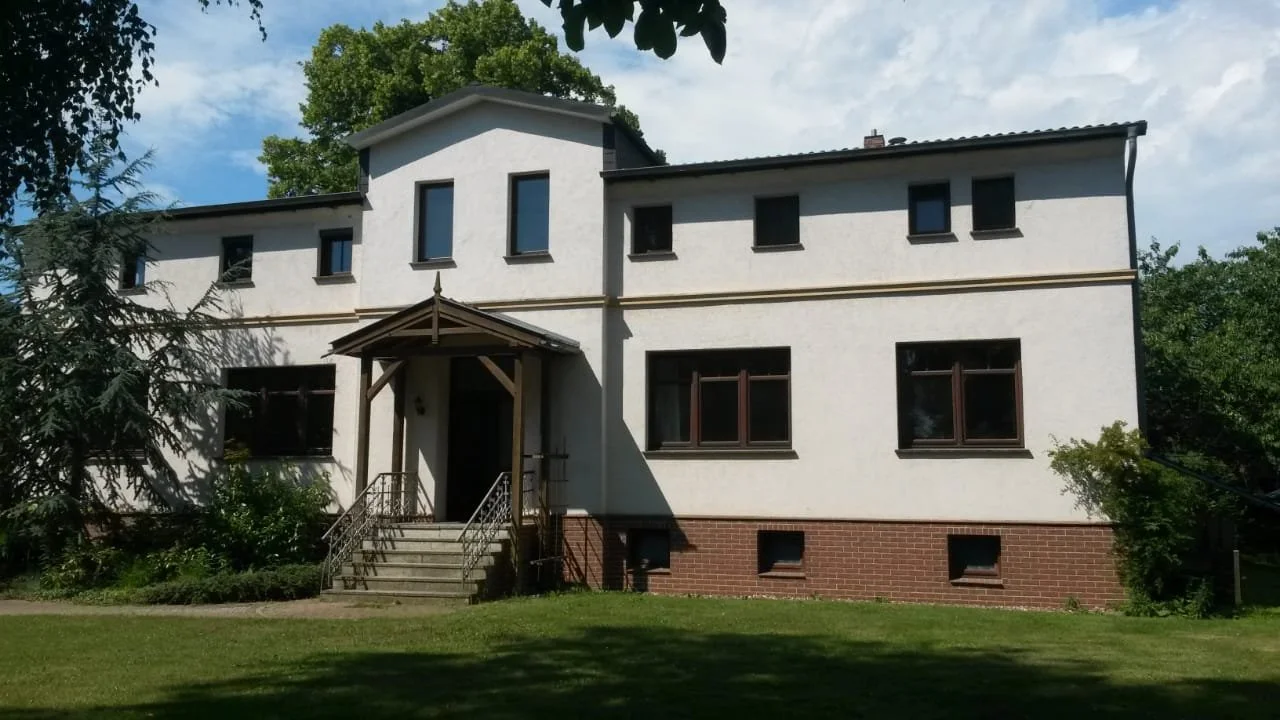 Ein zweistöckiges Haus mit weißer Fassade, braunen Fenstern und einem kleinen Vordach über der Eingangstür, umgeben von Bäumen und Rasen.