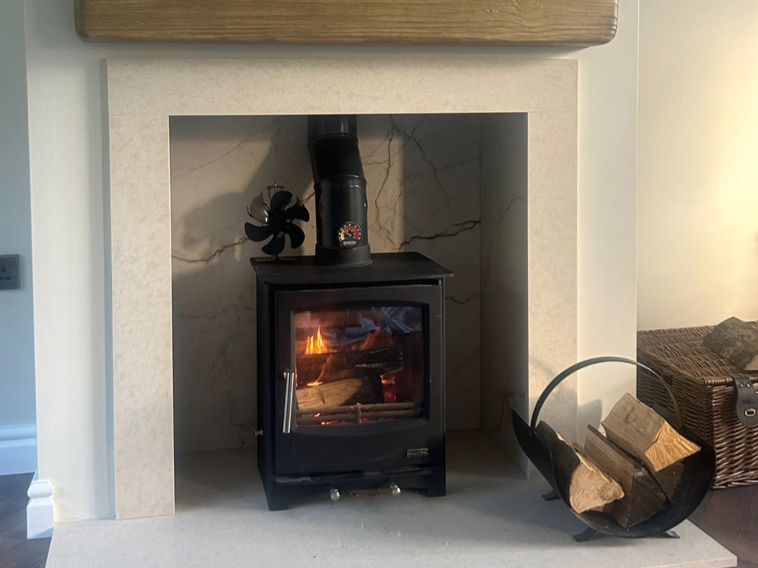 A wood-burning stove inside a fireplace with a small fire burning, black chimney pipe on top, a small black fan to the left, and a wicker basket with extra firewood to the right.