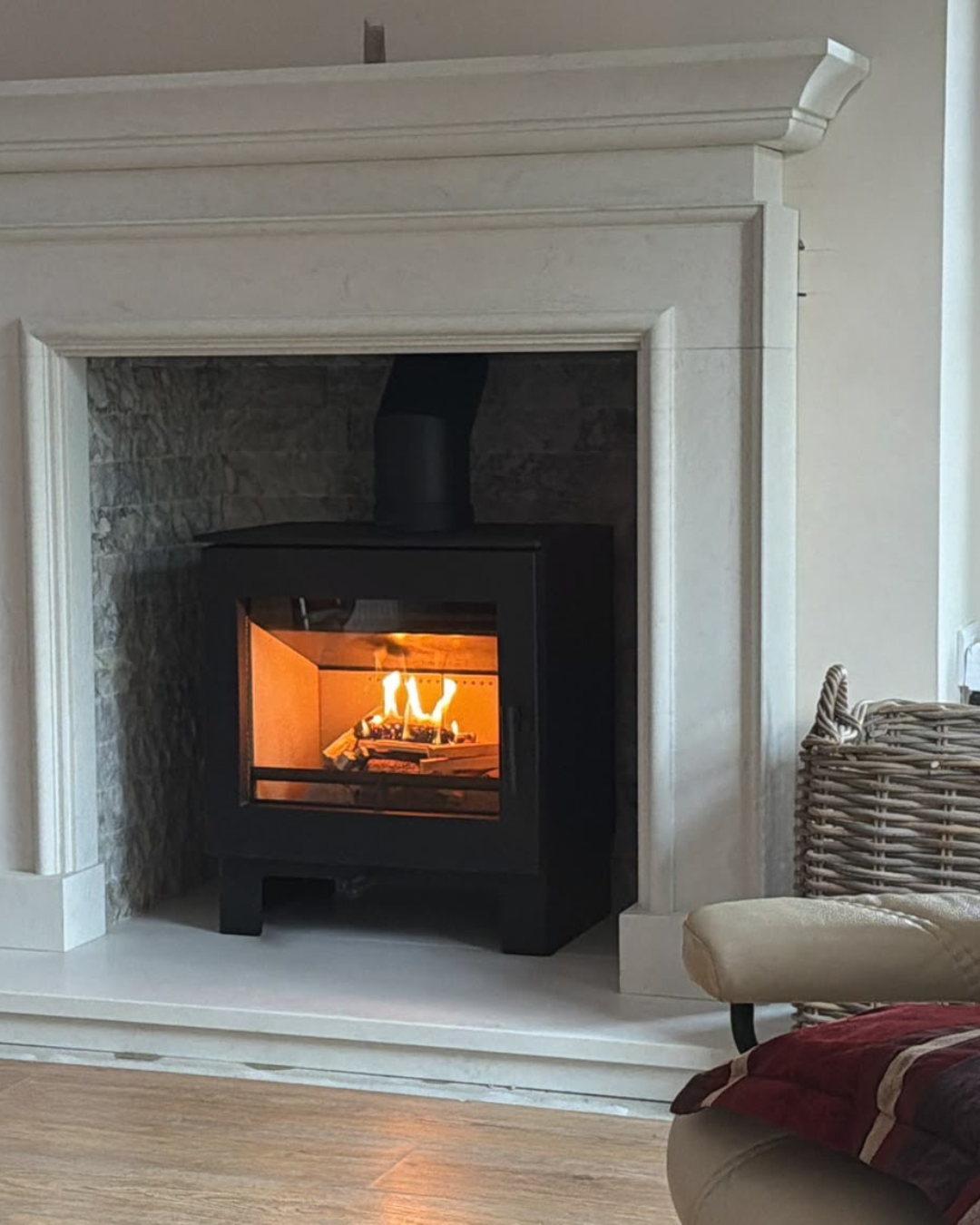 A black wood stove burning inside a fireplace with a stone interior, surrounded by a white mantel and hearth, with a wicker basket and cream-colored chair nearby.