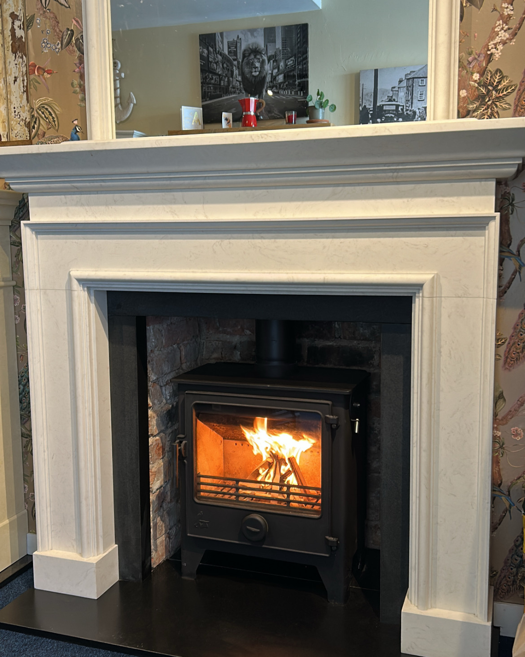A fireplace with a lit wood-burning stove inside a white mantel, with a mirror above reflecting part of the room, including framed black-and-white photos, a small potted plant, and decorative items.
