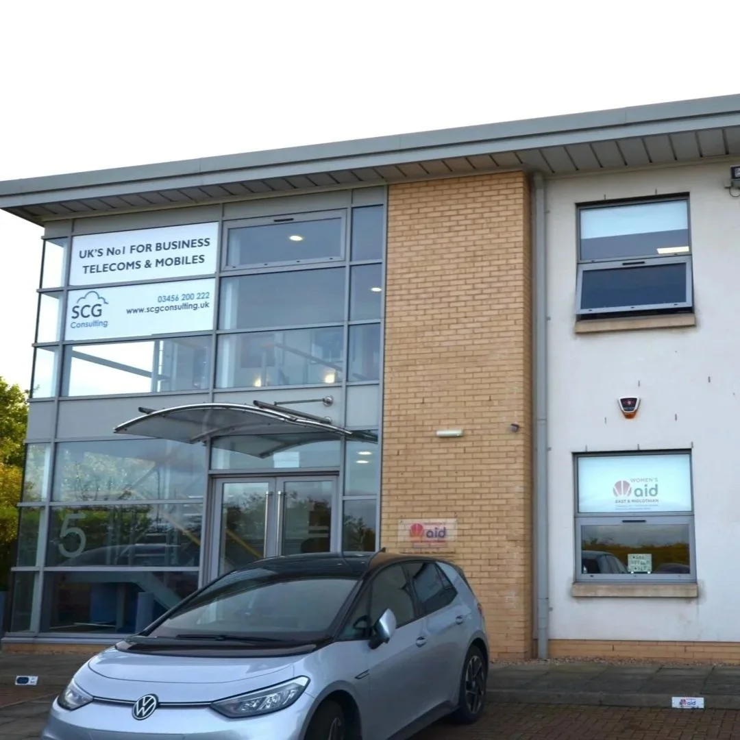 Front of Macmerry Business Park with a glass entrance, brick and white walls, and signage for 'SCG Consulting' and 'Women's Aid.' A silver Volkswagen car is parked outside.