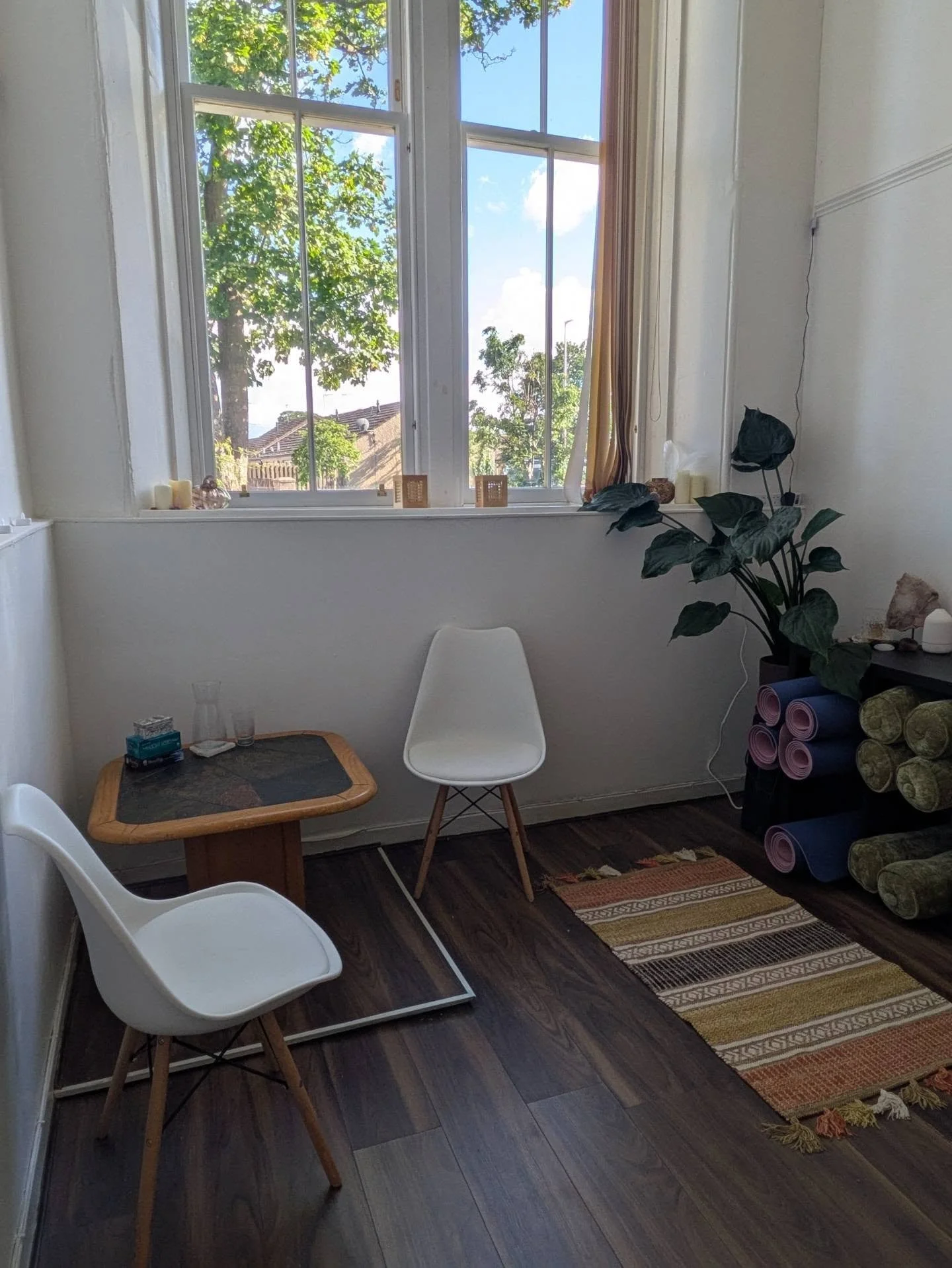 A cozy indoor space with a large window, white walls, and wooden flooring. The window offers a view of trees and a house outside. Inside, there are two white chairs, a small wooden table with a glass, tissues, and other small items. A tall plant with large dark green leaves is on the right, next to a rack holding rolled-up yoga mats. A colorful woven rug lies on the floor.