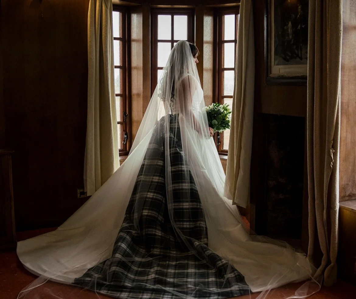A bride standing by a window, wearing a wedding dress with a plaid skirt underneath and a long veil, holding a bouquet of flowers.