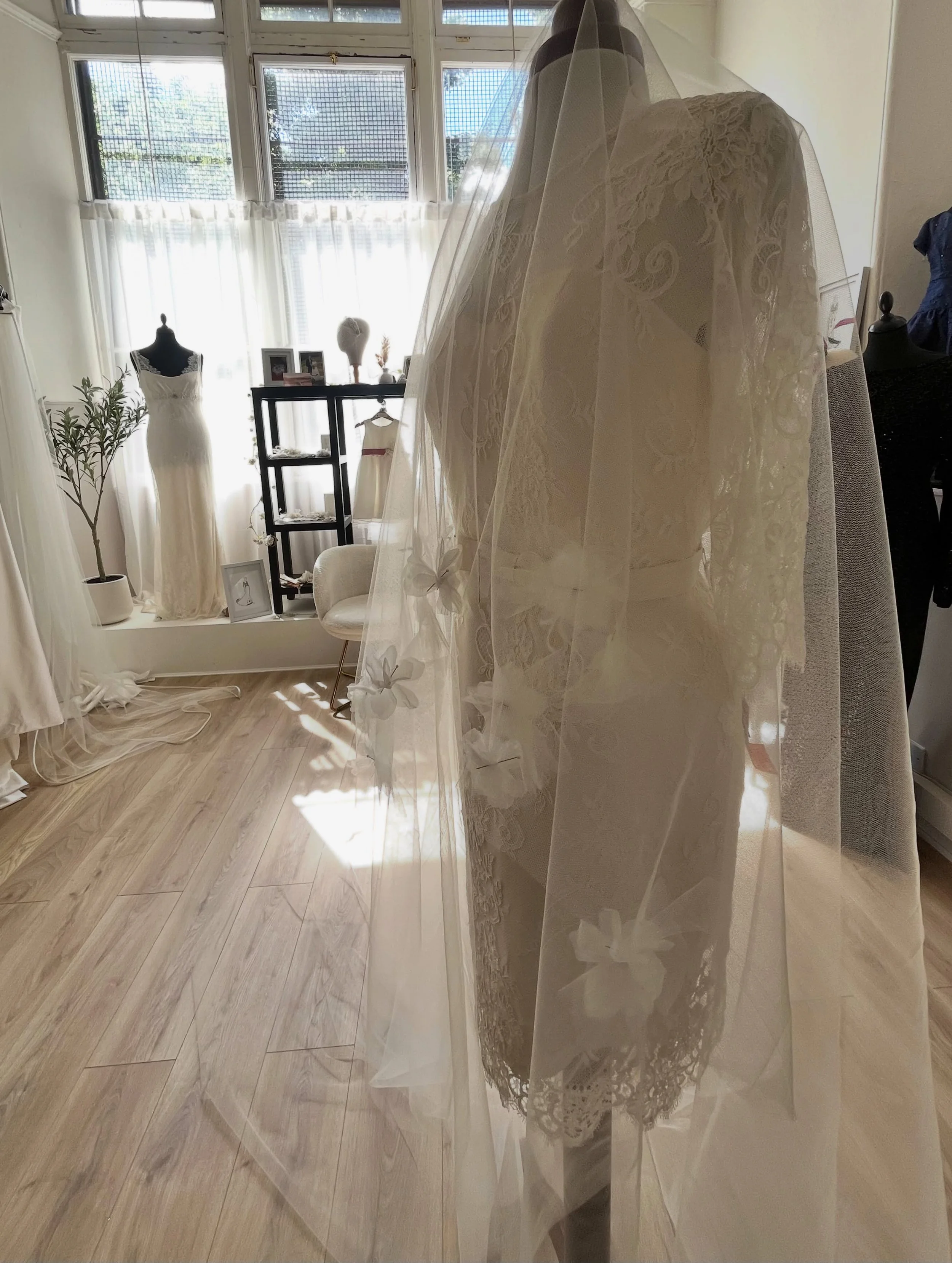 Interior view of a tailor, featuring mannequins dressed in wedding gowns, a small potted plant, framed photographs, and decorations on a shelf near large window with white curtains letting in natural light.