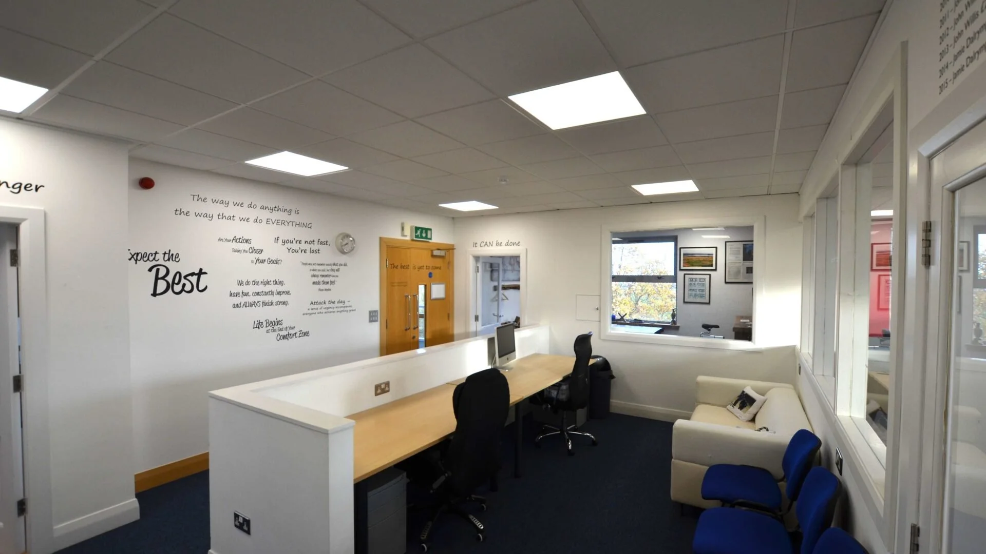 An office room with white walls decorated with motivational quotes, a light-colored wooden desk with a computer monitor, black chairs, and a white sofa with a pillow. There are large windows on the right side and a door with a glass window on the back wall. The ceiling has white tiles with fluorescent lighting.