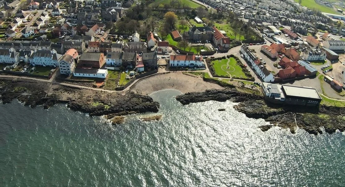 Aerial view of Cockenzie, taken by Boatshore Cottage, showing houses, a small beach, and a rocky shoreline along the ocean.