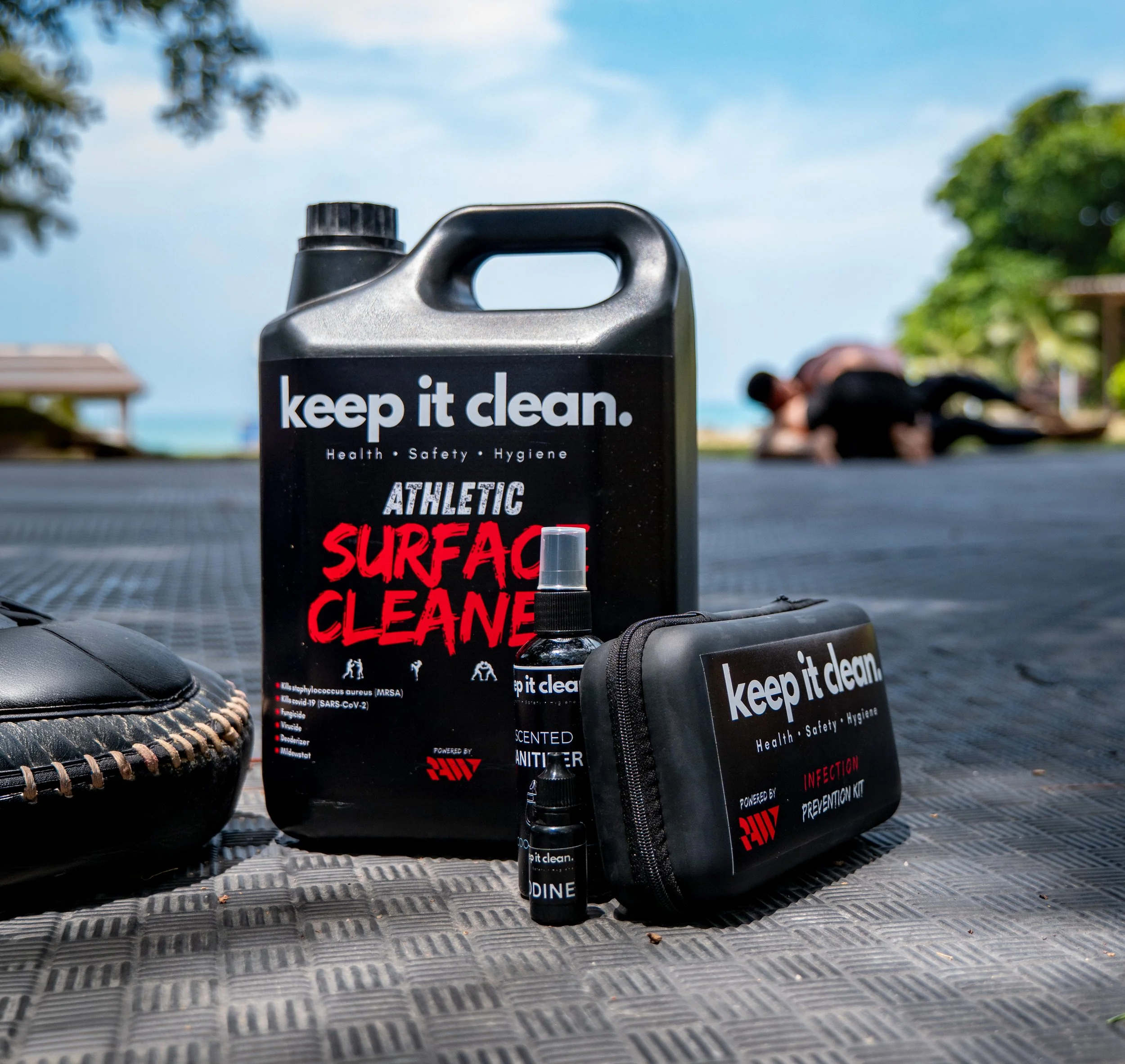 Schwarze Flasche mit Weißschrift, die sagt 'keep it clean', auf einem rutschfesten schwarzen Boden, im Hintergrund ein grüner Baum, blauer Himmel und eine Person, die im Fitnessstudio trainiert.