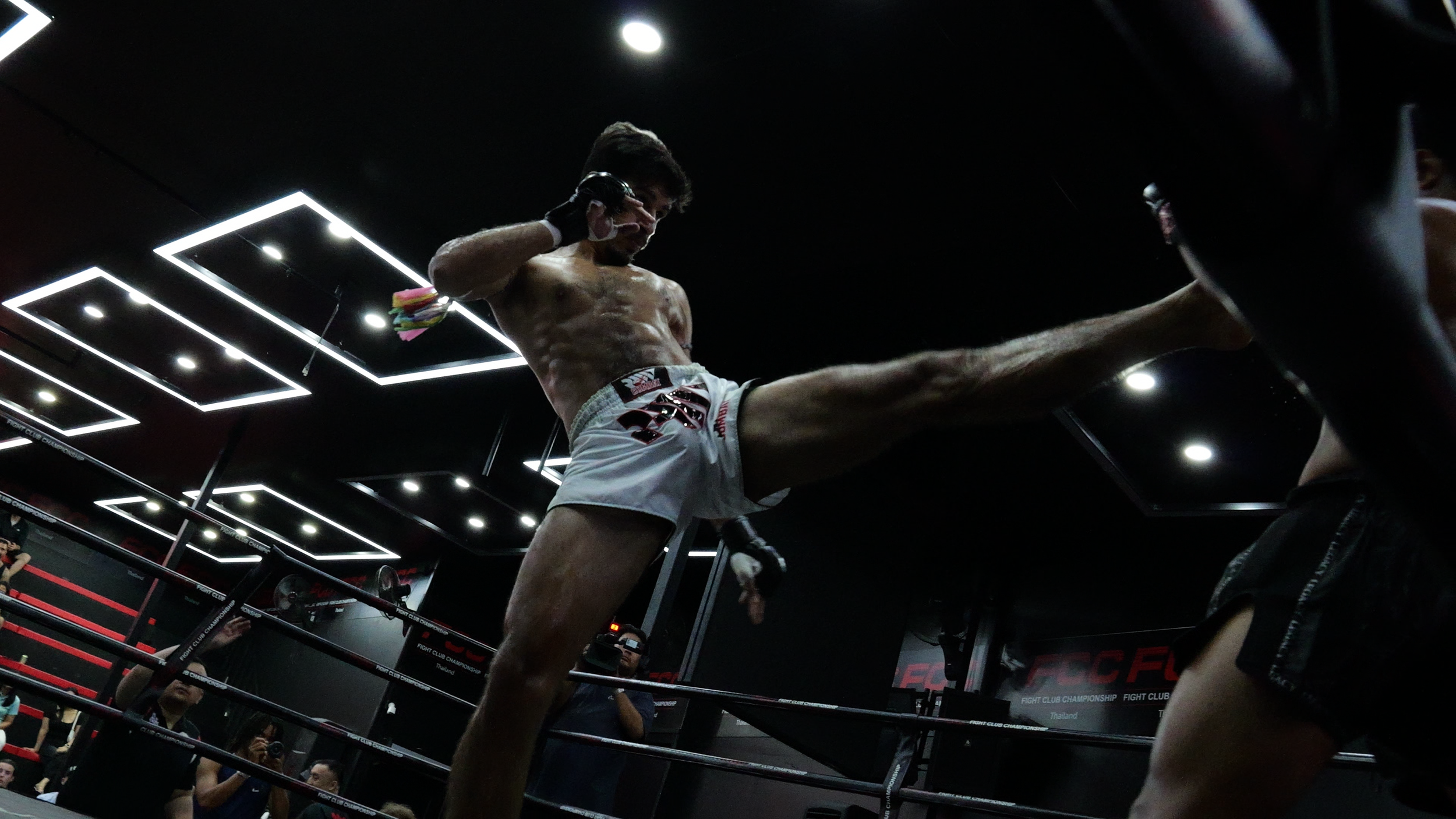 Mann trägt Boxhandschuhe und Shorts und führt einen Tritt im Muay-Thai-Kampfring aus, während er auf einen Gegner zielt, im dunklen Raum mit LED-Beleuchtung.