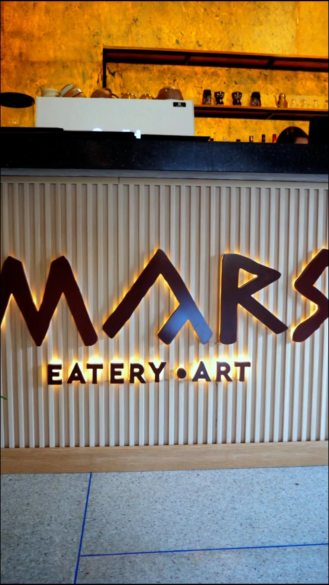 Blick auf eine Theke in einem Restaurant mit einem beleuchteten Wandlogo, das 'WOKS' und darunter 'EATERY · ART' zeigt.