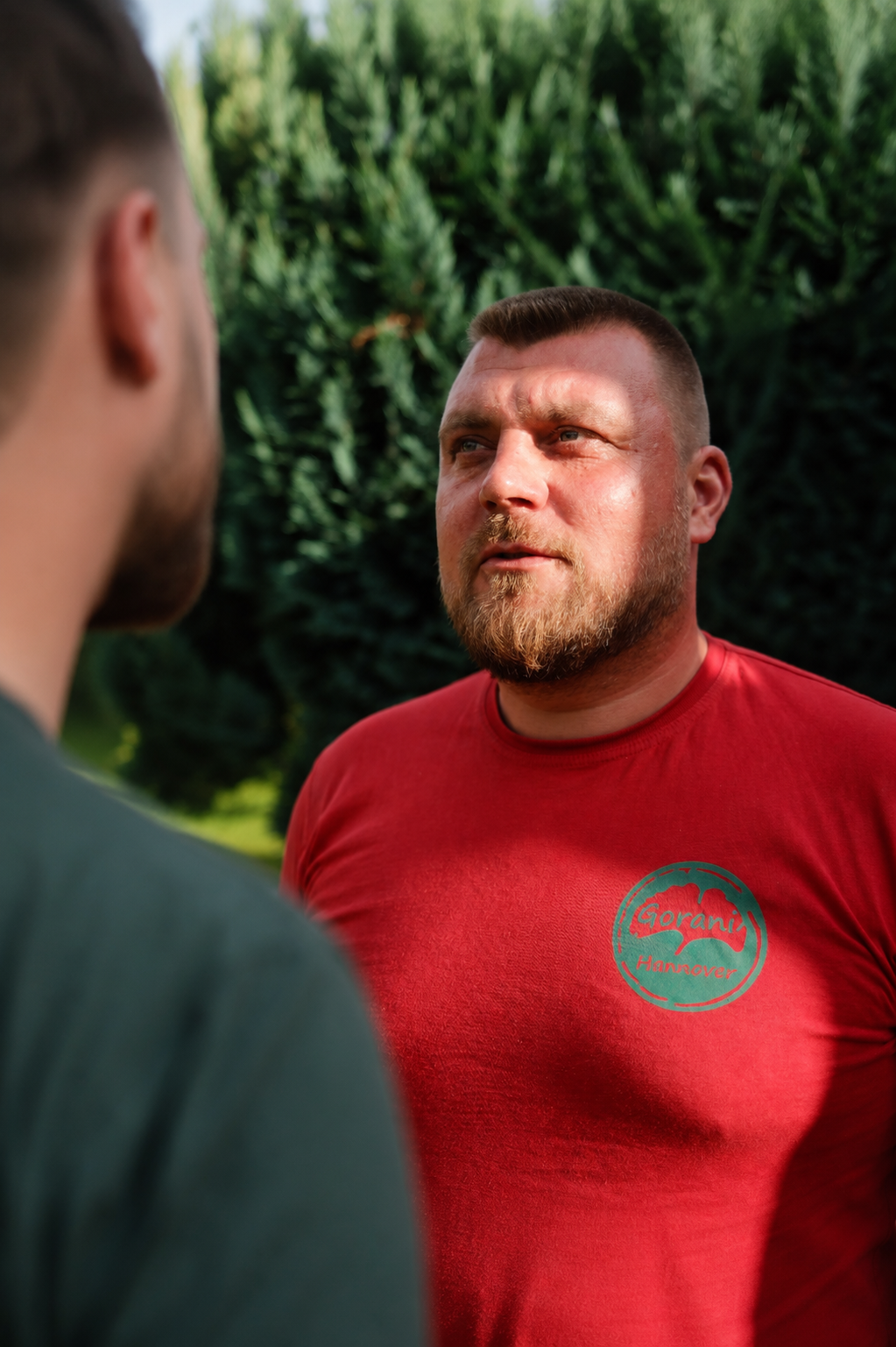 Zwei Männer stehen sich draußen gegenüber, im Fokus ist ein Mann mit rotem T-Shirt, während ein anderer Mann nur halb sichtbar ist.