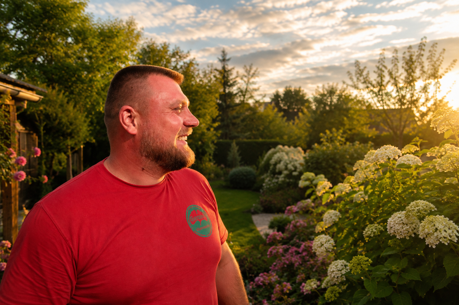 Ein lachender Mann in einem roten T-Shirt im Garten bei Sonnenuntergang, umgeben von blühenden Blumen und grünen Bäumen.