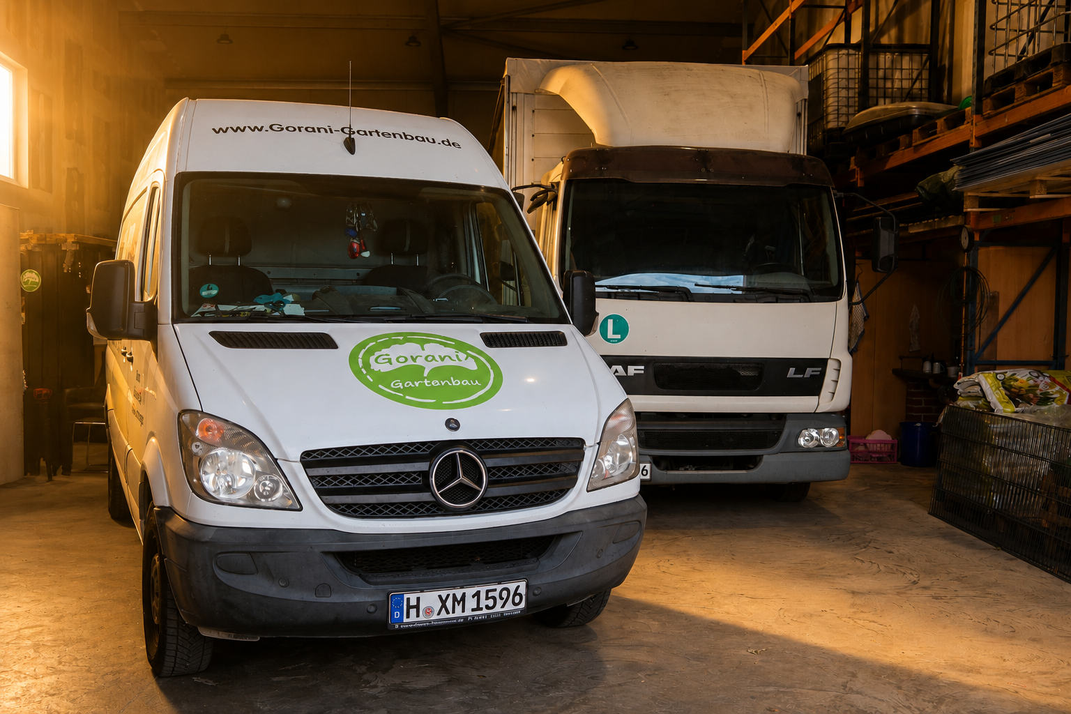 Zwei Firmenfahrzeuge in einem Lagerraum, ein Mercedes-Transporter mit 'Gorani Gartenbau' Logo vorne, dahinter ein LKW