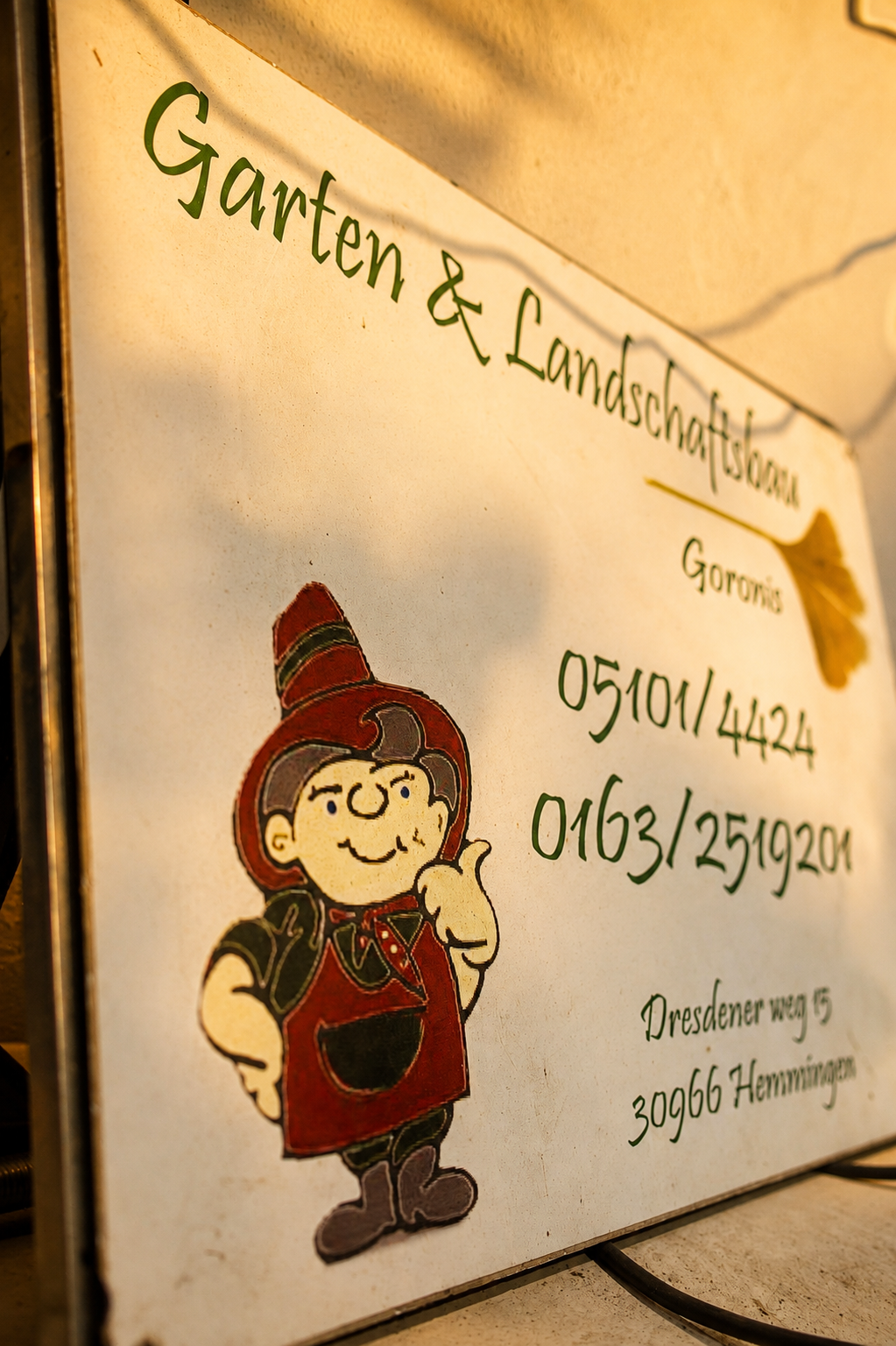 Werbetafel für Garten- und Landschaftsbau mit Kontaktinformationen und Cartoon-Illustration eines freundlichen Gartendieners.
