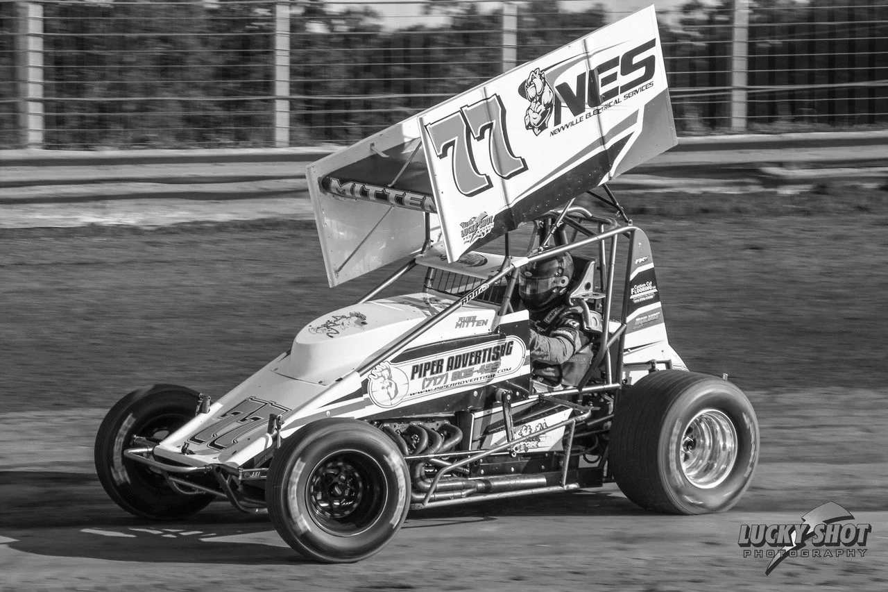 Russell Mitten in car. Black and white photo of a sprint car racing on a dirt track, with the driver wearing a helmet, and a large wing with the number 77 on top of the car.