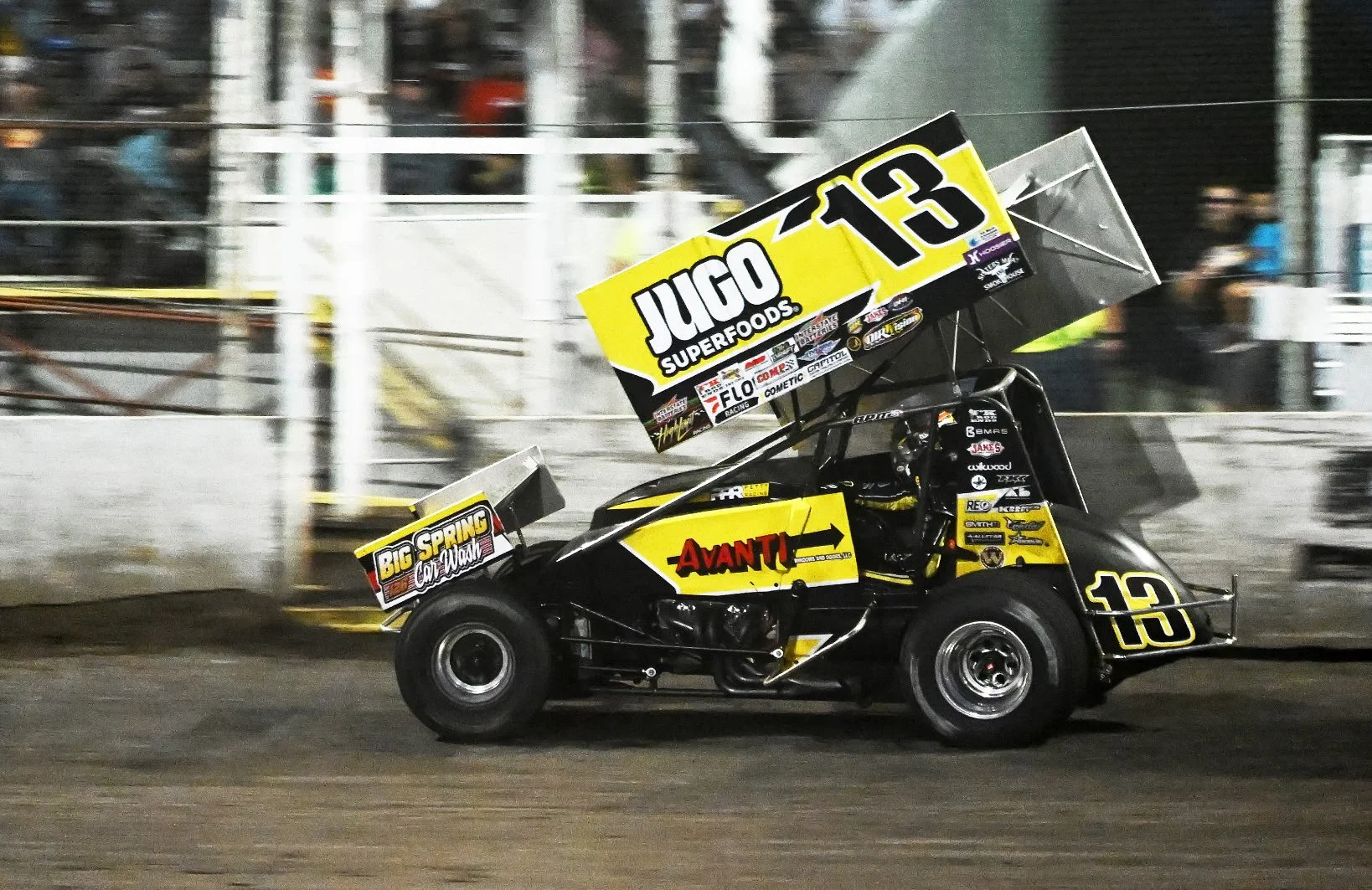 A sprint car racing on a dirt track with a yellow and black color scheme, featuring the number 13 and sponsor logos, in motion at night.