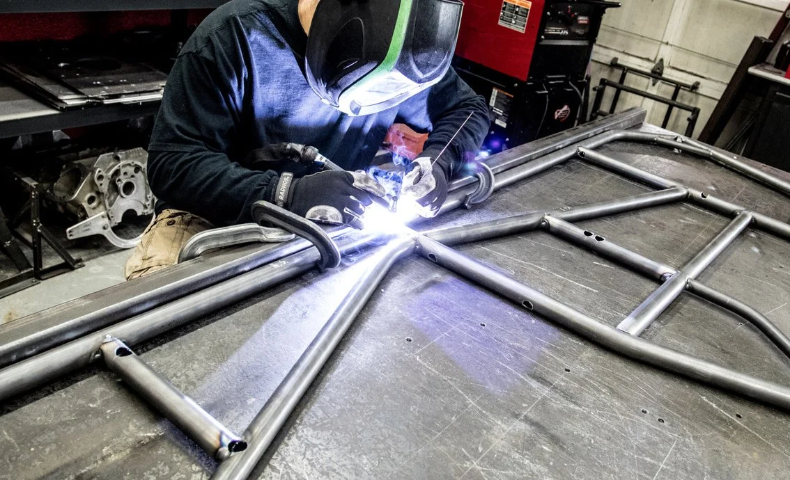 Person welding a metal framework in a workshop, wearing protective gear, including a welding helmet and gloves.