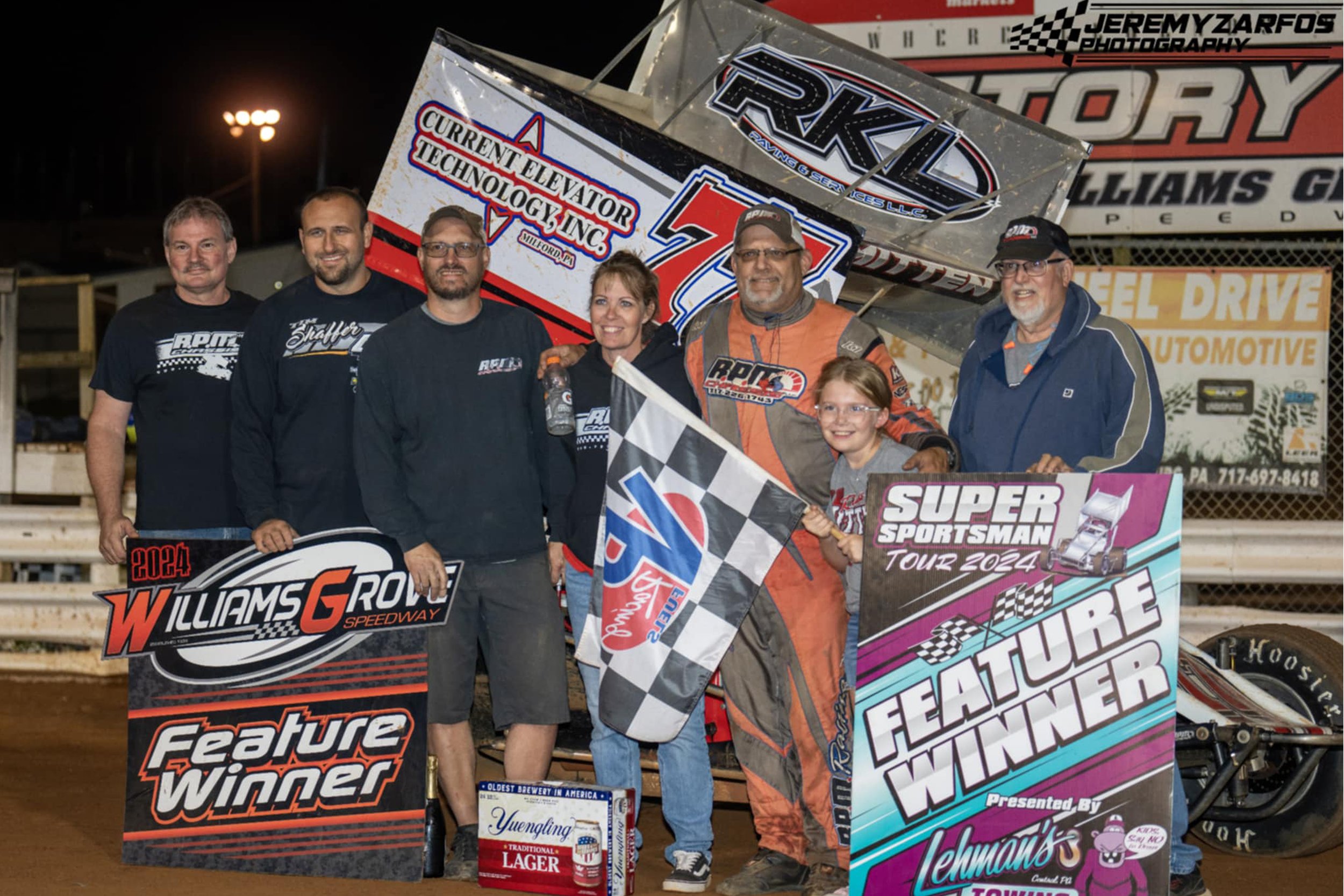 A group of people, including a racing driver in a racing suit, standing in front of a race car and holding a checkered flag at a race track at night. The scene celebrates a victory, with banners and signs related to the race and sponsor logos visible.