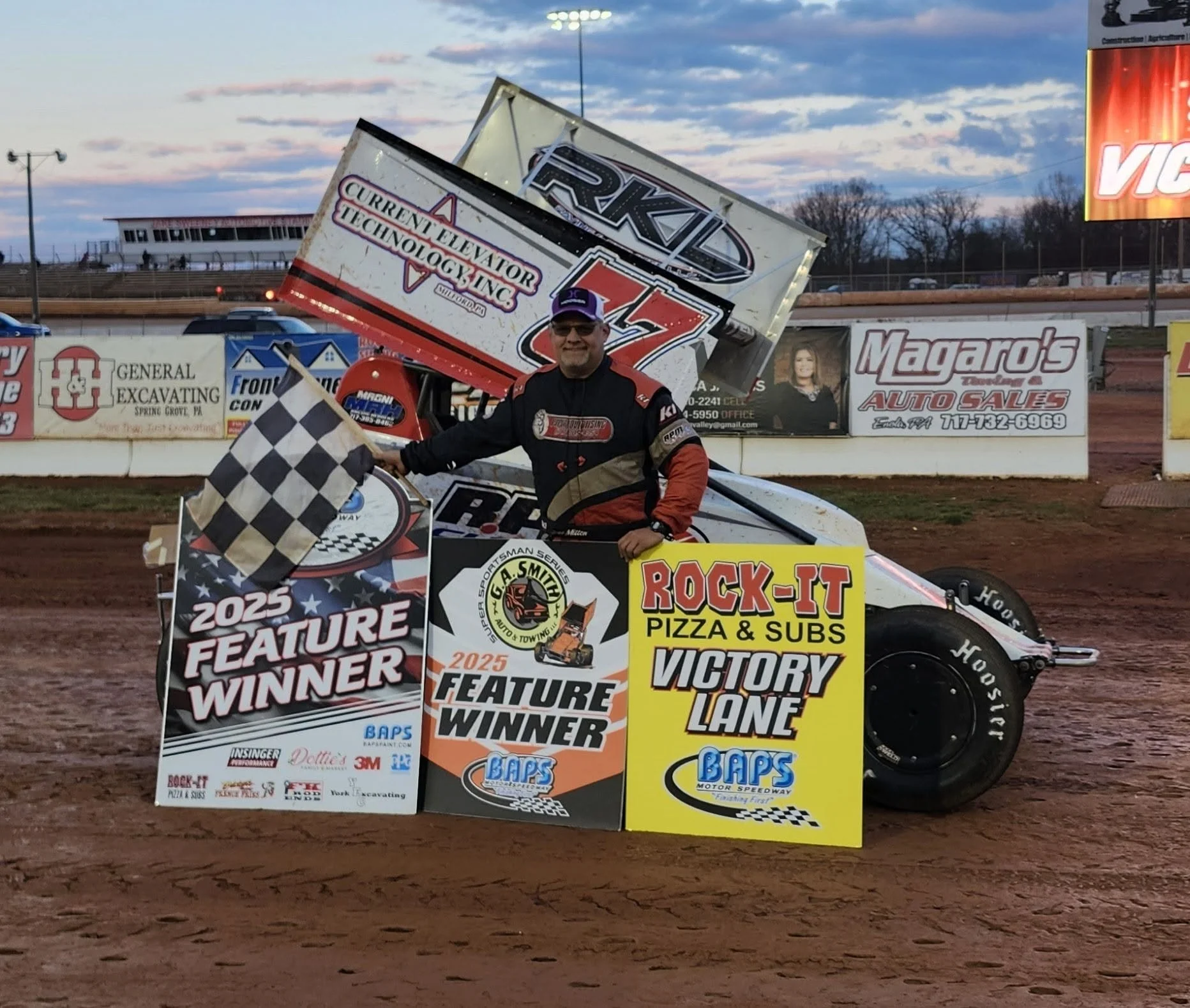 Race car driver in racing suit holding checkered flag, standing beside his race car at a dirt track, with victory signs and banners displaying race-related advertisements and sponsor logos in the background.