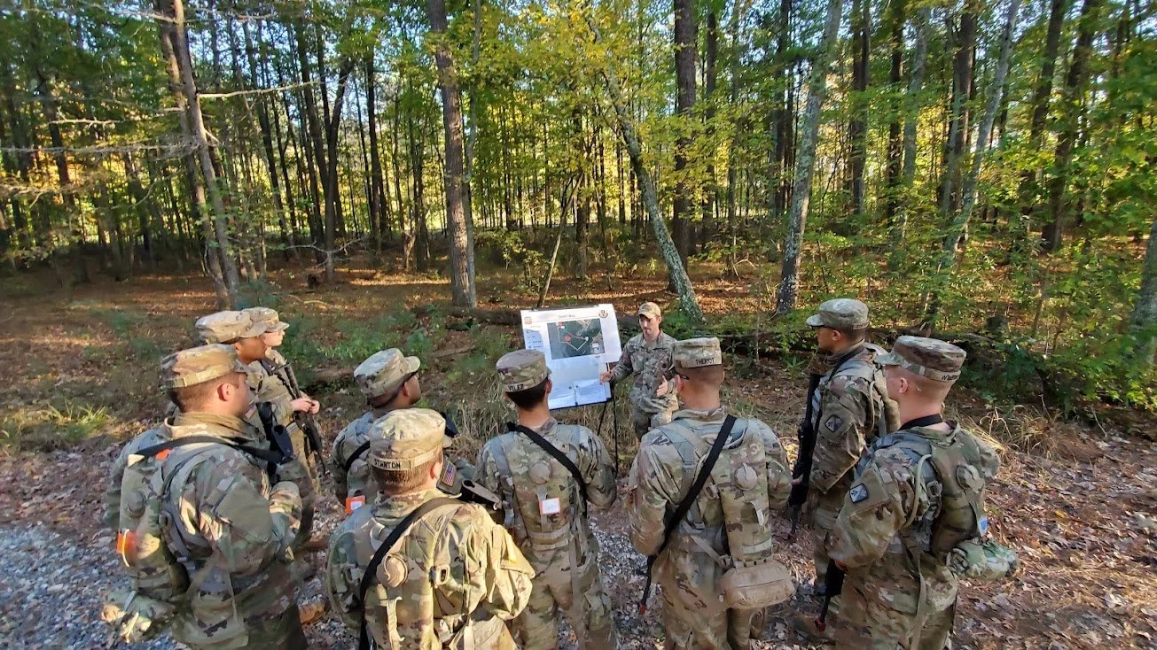 Military personnel in camouflage uniforms gathered in a forest, listening to a person at a presentation board.