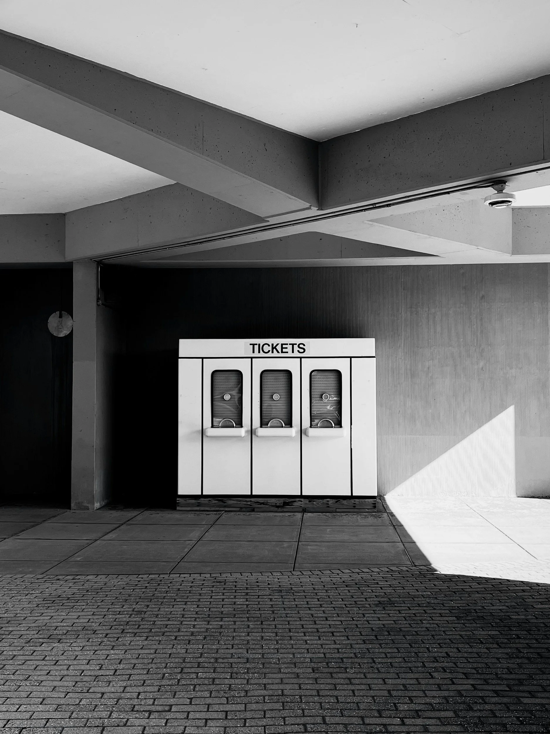 Black and white photo of an indoor space with a row of ticket vending machines against a wall, illuminated by sunlight, with tiled floor and a ceiling with visible concrete beams.