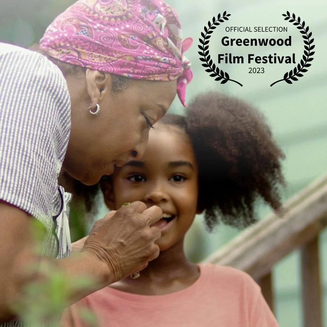 An older woman wearing a pink headscarf shows something to a young girl who has curly hair, both smiling, with a green background and a wooden railing. There is a black laurel wreath with the text 'Official Selection Greenwood Film Festival 2023' in the top right corner.
