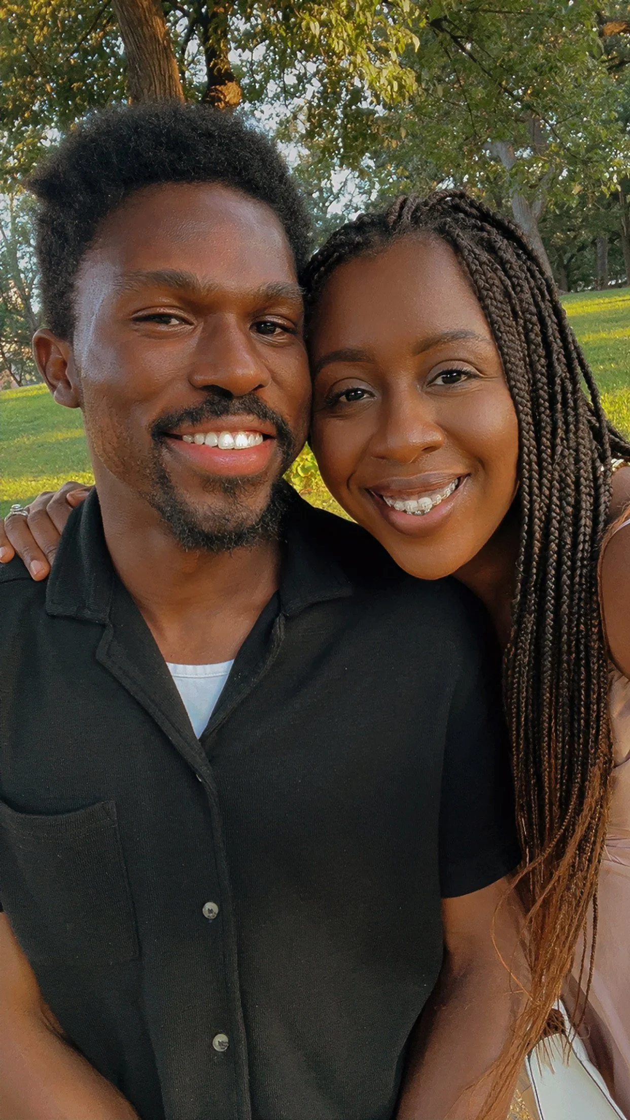 A happy couple taking a selfie outdoors in a park with trees and grass in the background. The man has short curly hair and a beard, wearing a black shirt, and the woman has long braided hair, smiling with braces.