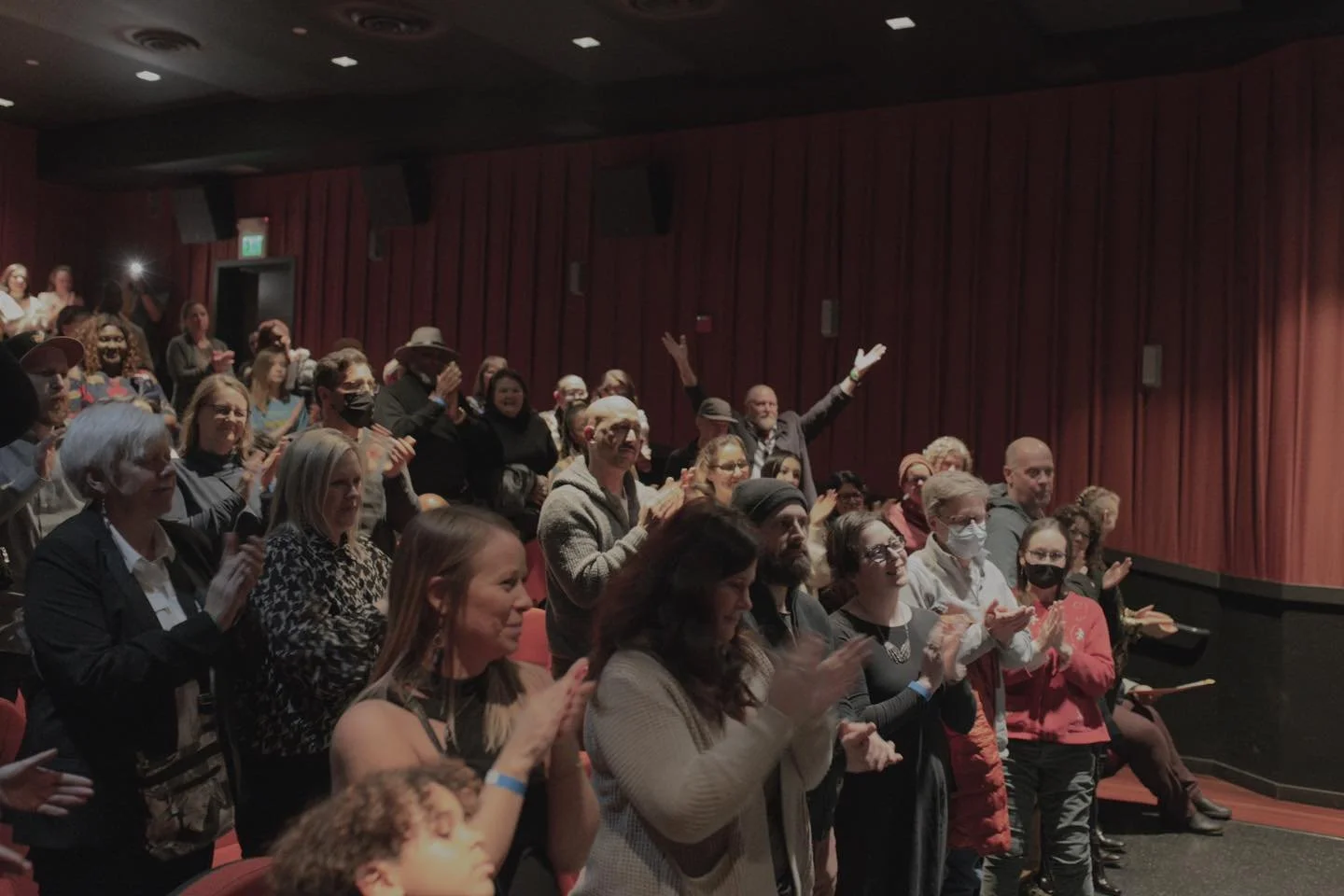 Audience standing and clapping in a theater or performance venue with red curtains on the stage.
