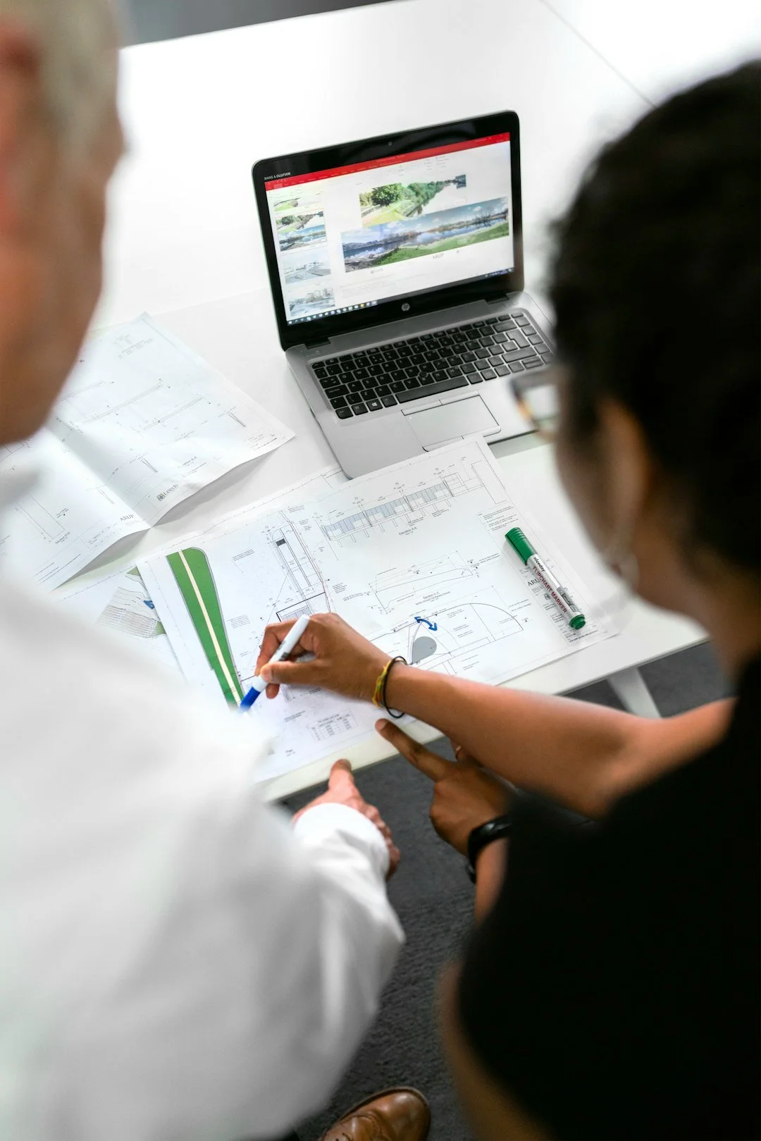 Two people working at a desk with architectural blueprints, a marker, and a laptop displaying landscape photos.