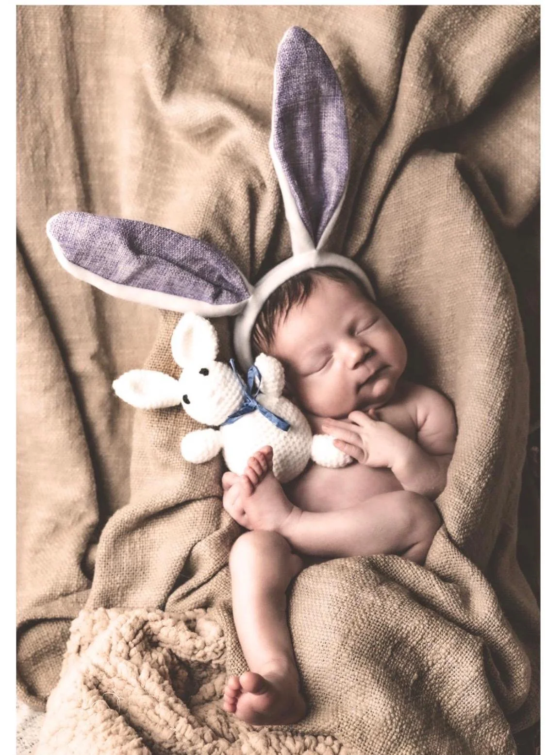 Un bambino neonato dorme avvolto in una coperta, indossa una fascia con orecchie da coniglio e tiene un coniglietto di peluche. Trasmette dolcezza e tenerezza.