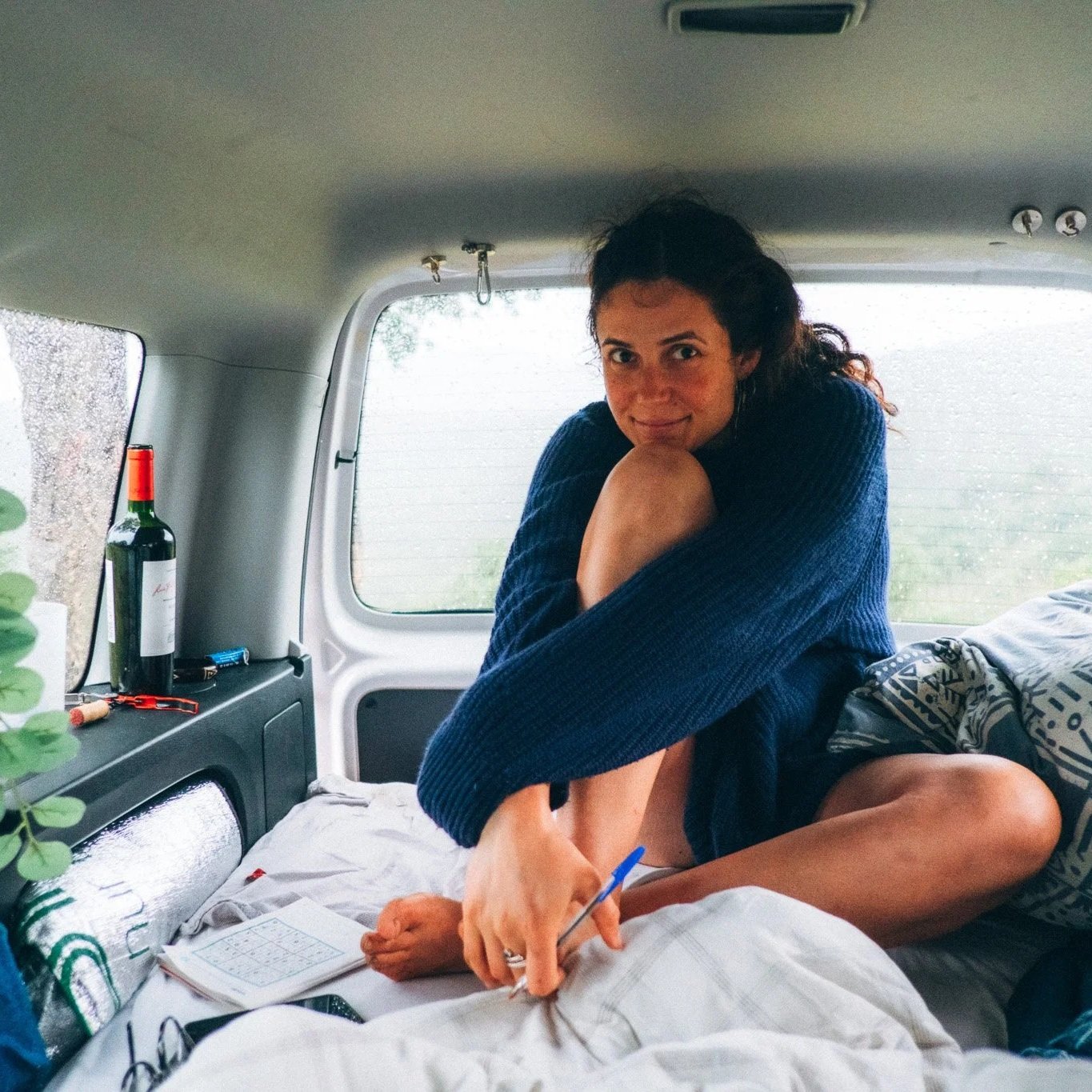 Eine junge Frau sitzt in einem Van und schreibt in ein Notizbuch, während sie sich umarmt. Der Van ist innen mit einem Bett ausgestattet, auf dem sie sitzt, mit einem Kissen und Decken. Auf einer Ablage im Hintergrund stehen eine Flasche Wein, eine Z