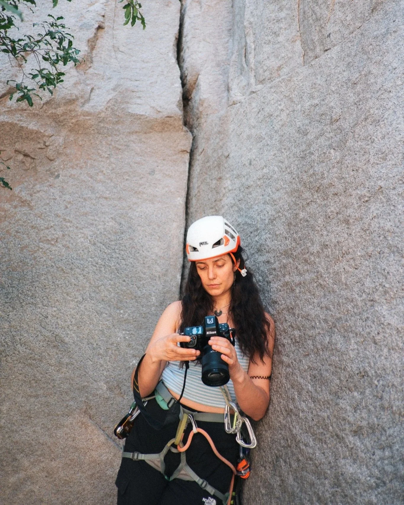 Junge Frau mit Kletterausrüstung und Helm, die einen Fotoapparat hält, steht vor einer Felswand.
