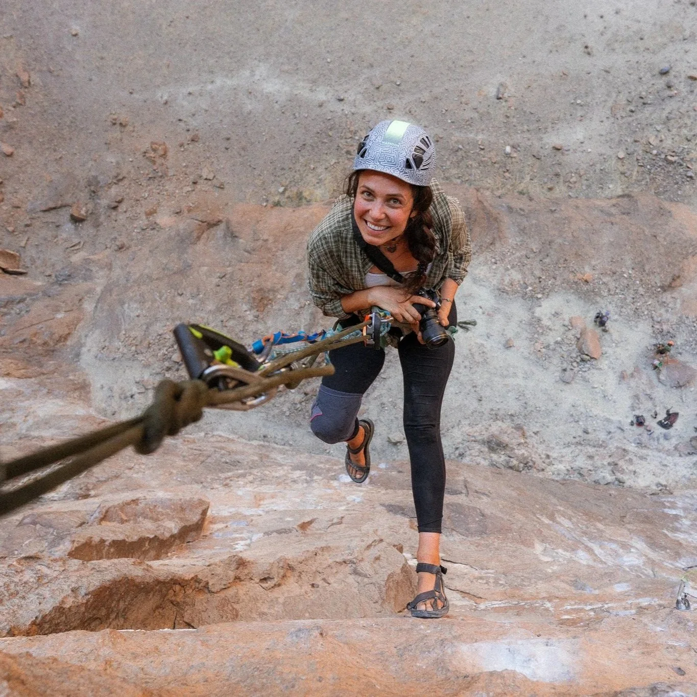 Eine Frau klettert an einer Felswand im Sandstein-Klettergebiet, trägt einen Kletterhelm und fotografiert sich selbst mit einer Kamera, während sie nach oben klettert.
