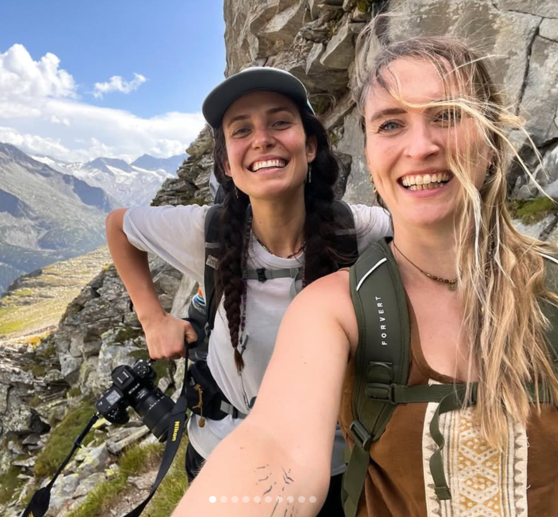 Zwei lachende Frauen beim Wandern in den Bergen mit Rucksack, vor einer Felswand, umgeben von Bergketten und Wolken