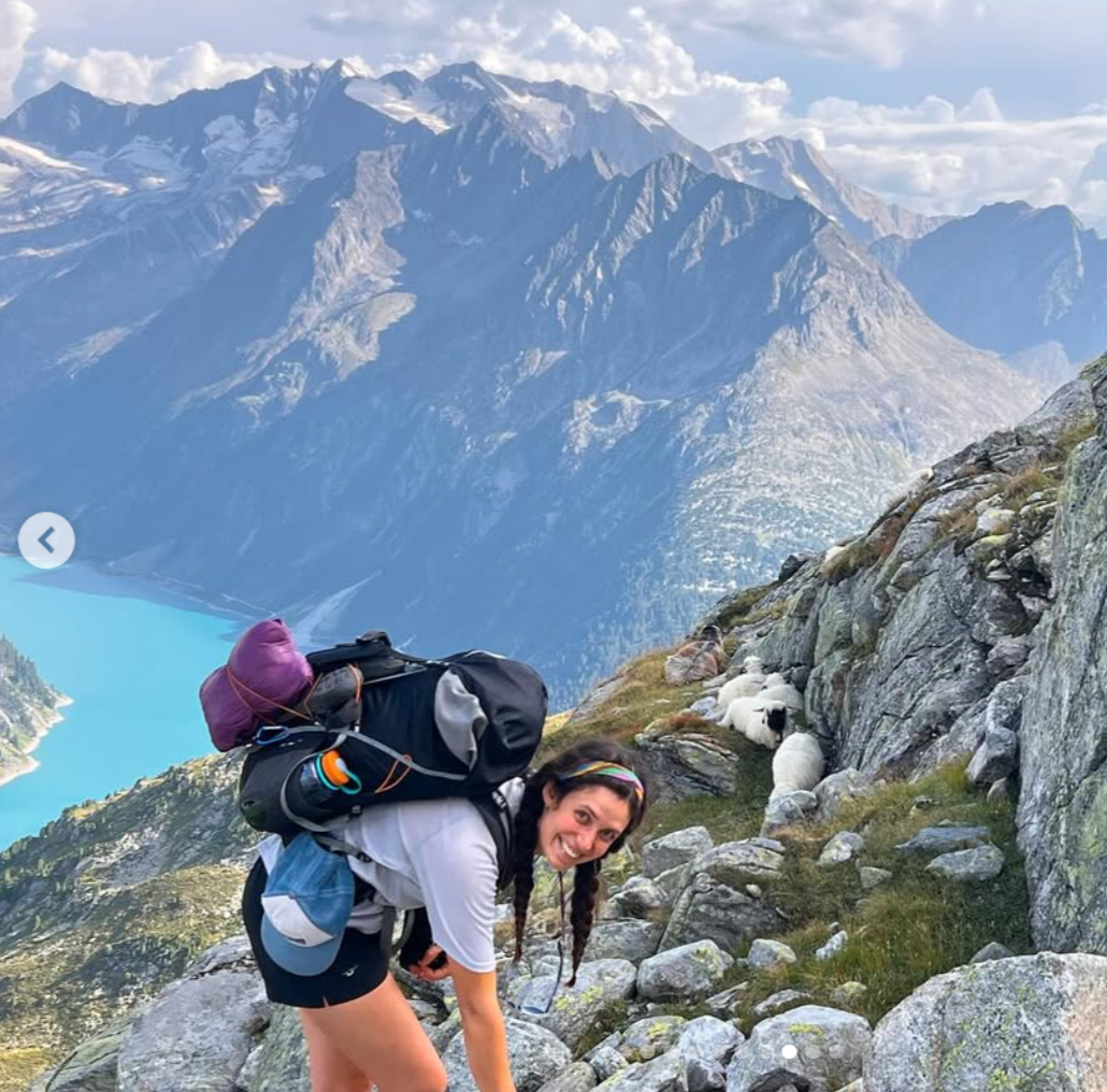 Eine Frau beim Klettern am Berg, umgeben von Felsen, mit einer Berglandschaft im Hintergrund, darunter ein See, hohe Berge und Wolken am Himmel.