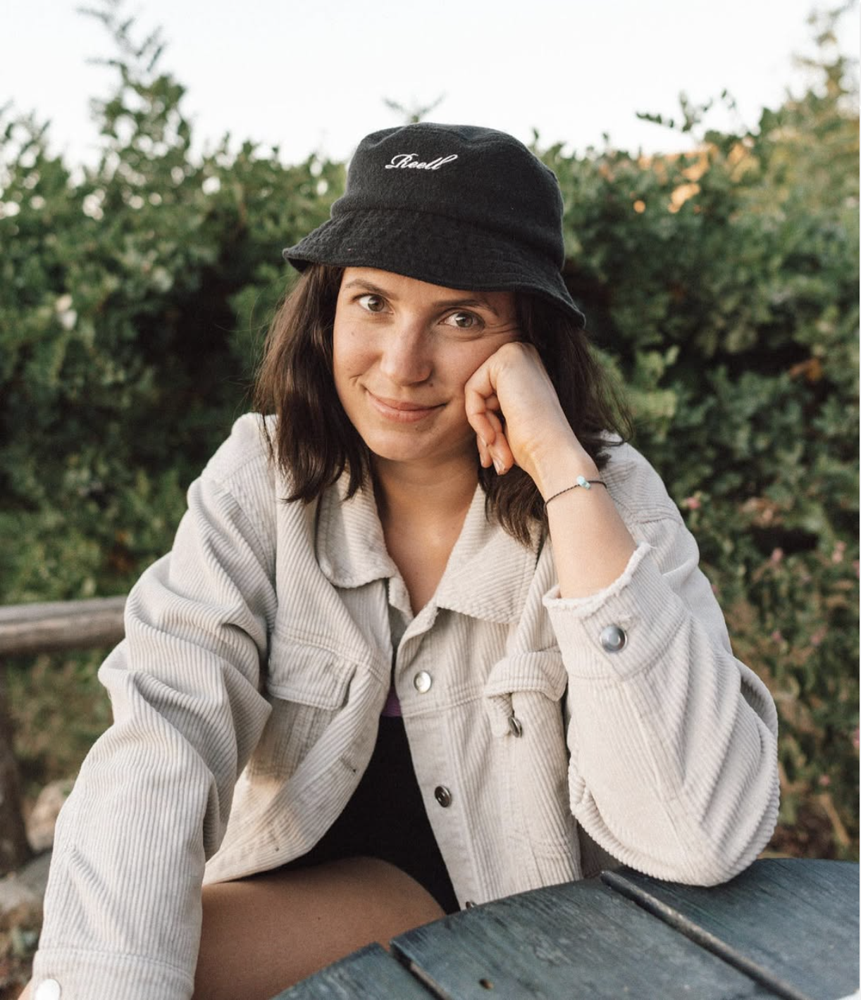 Eine junge Lifestyle Fotografin mit dunklen Haaren, die eine schwarze Bucket-Hat trägt, sitzt draußen vor einer grünen Hecke und lächelt freundlich in die Kamera.