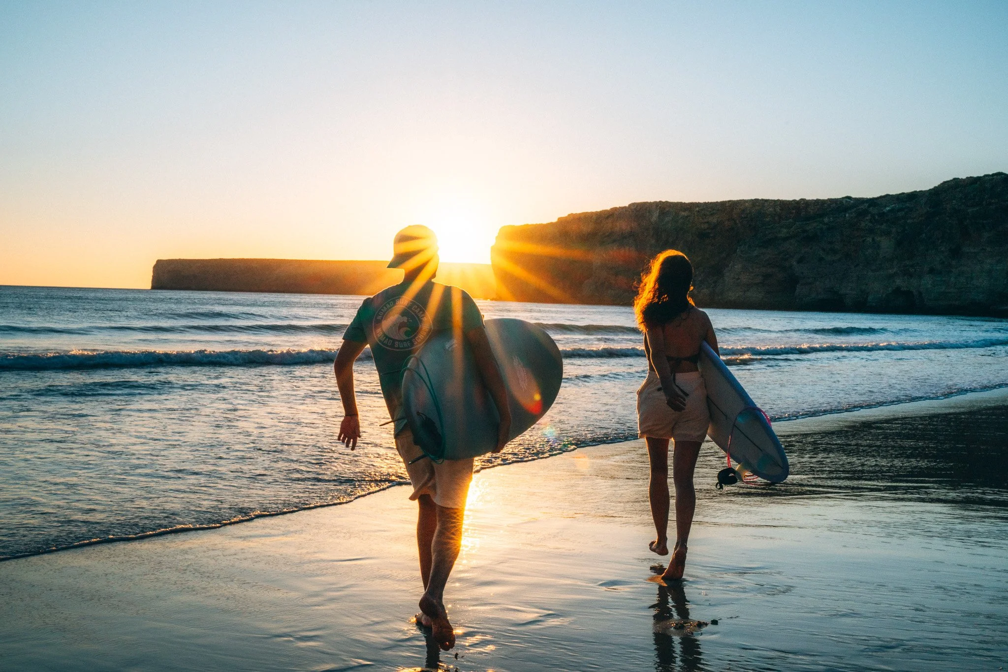 Lifestyle photographyzweier Surfer die mit Surfbrettern am Strand bei Sonnenuntergang ins Wasser rennen.