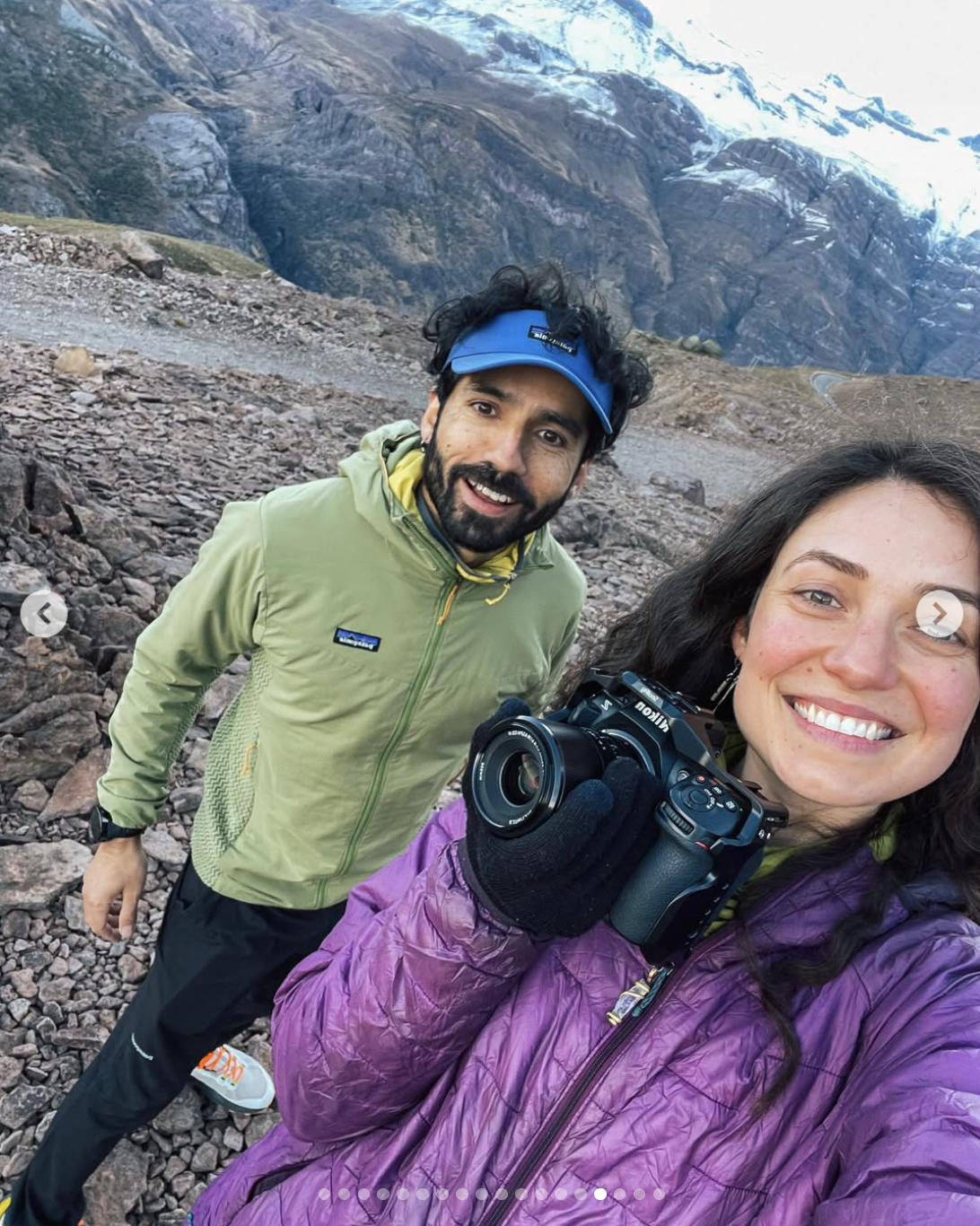 Ein lachendes Paar macht ein Selfie vor einer Bergkulisse mit Schnee auf den Gipfeln, beide tragen Outdoor-Kleidung für eine Bergwanderung.