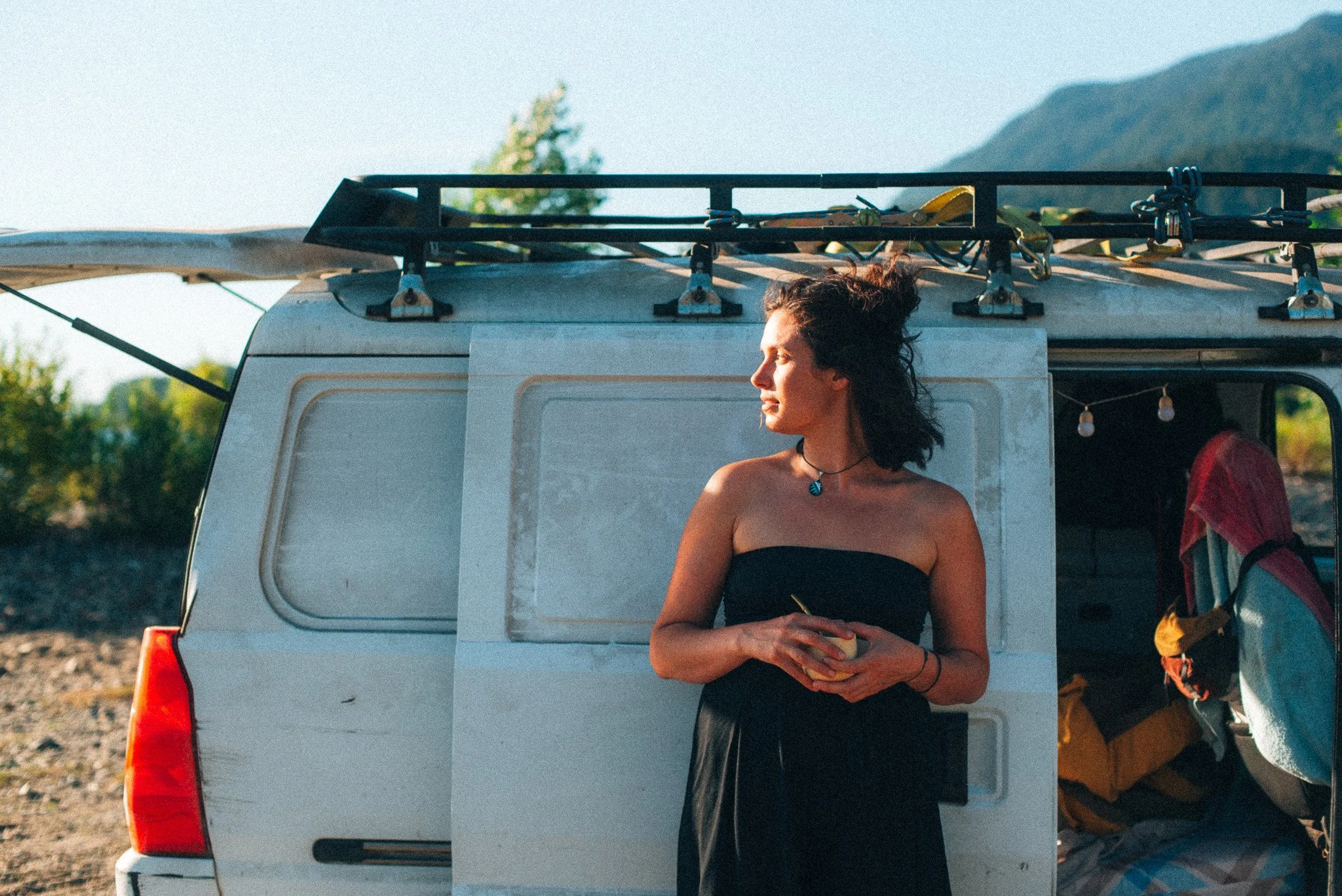 Junge Frau steht neben einem weißen Wohnmobil mit aufgesetztem Dachgepäckträger, Sonnenlicht, Berge im Hintergrund, trägt ein Schwarzes Oberteil, Halskette, hält eine Tasse in den Händen.