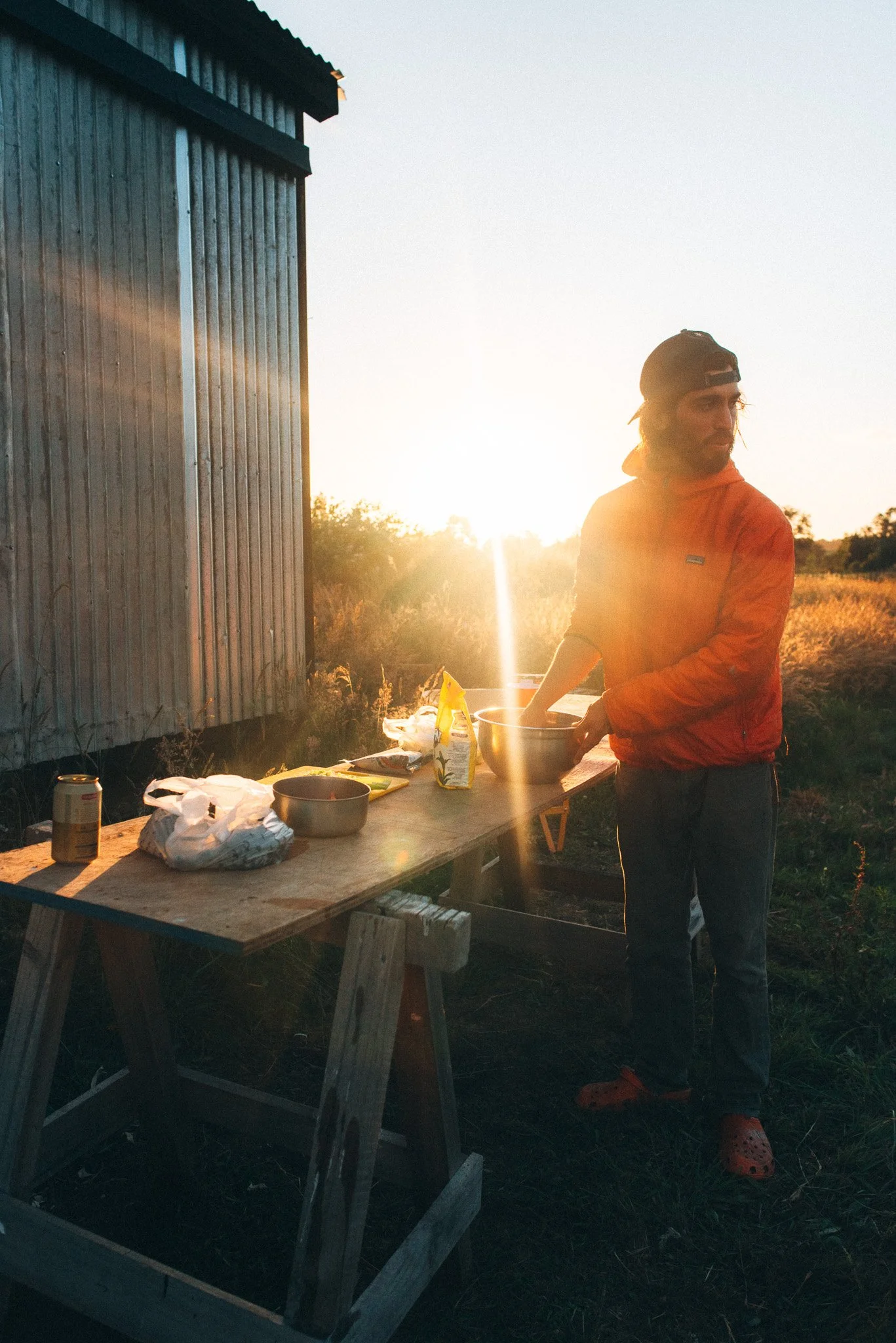 A man wearing an orange jacket, black baseball cap, and red Crocs standing outdoors at sunset, preparing food on a makeshift wooden table with various food items and containers, next to a metal shed in a rural setting.